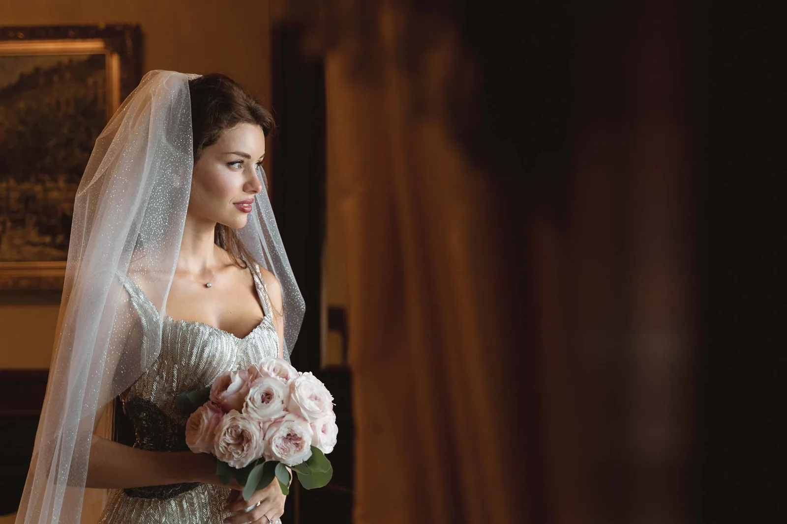 La sposa con il velo e il bouquet di rose rosa, nella penombra della camera