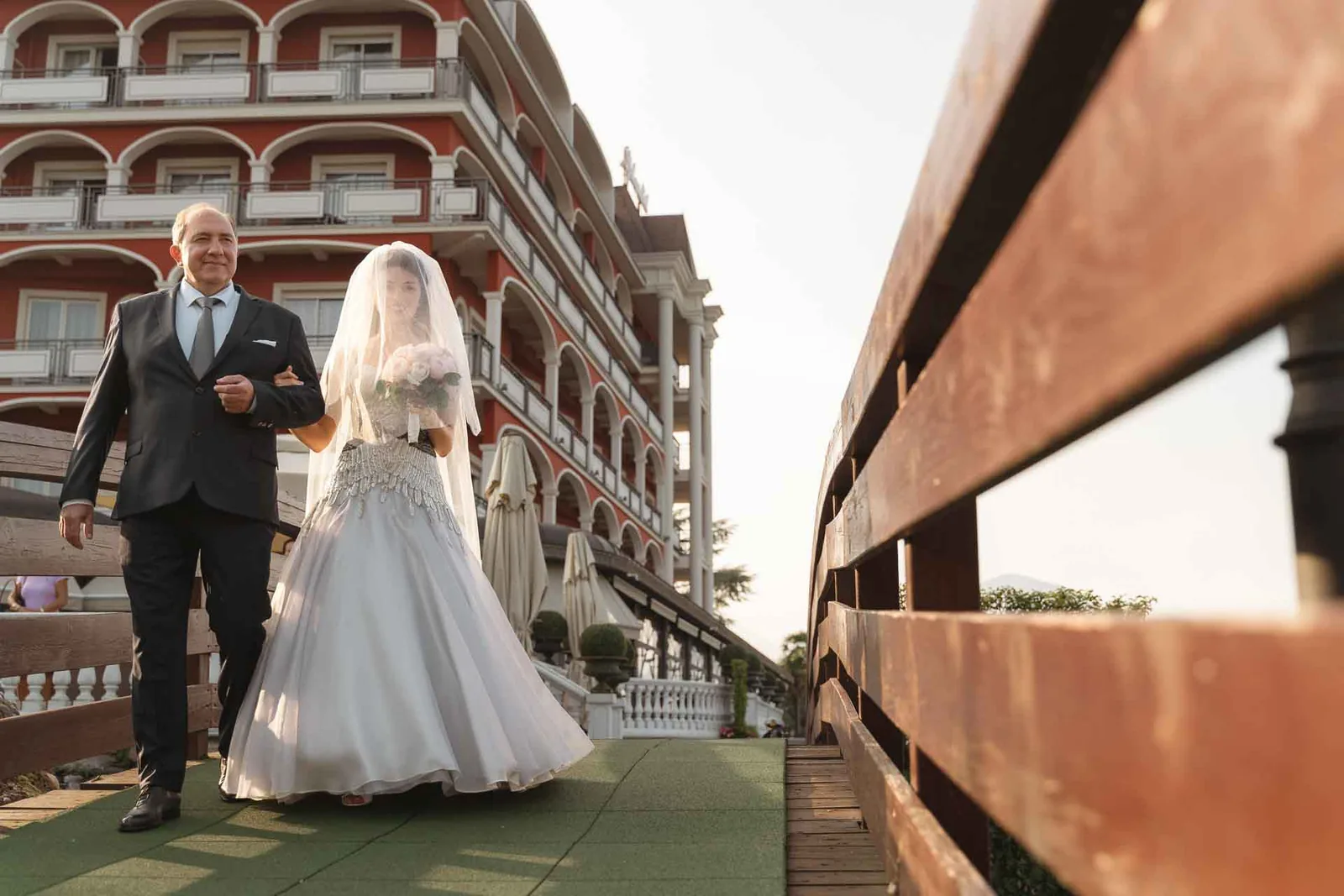 La sposa attraversa il ponte di legno sottobraccio, verso la cerimonia