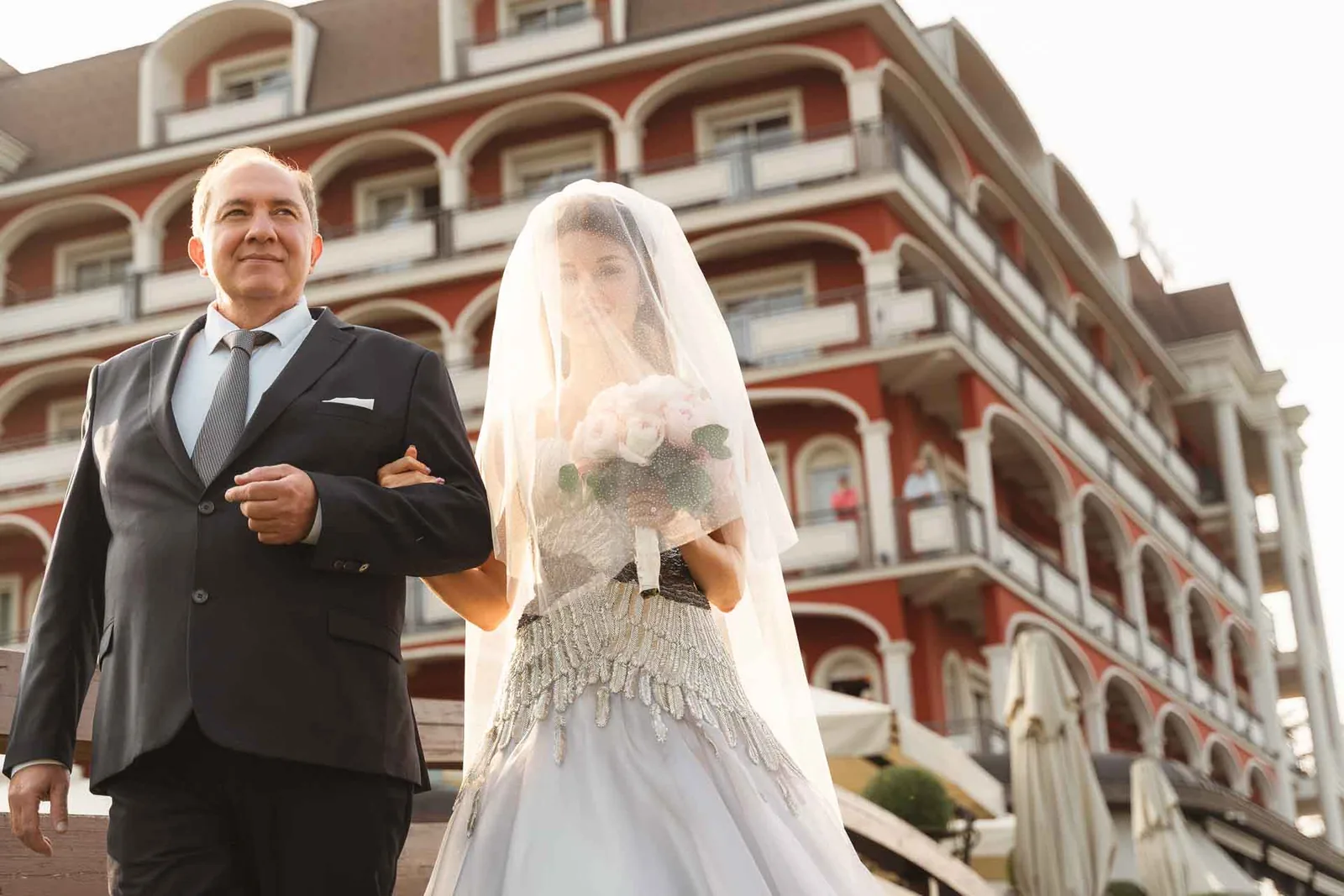 La sposa con il velo sul viso, sottobraccio, davanti alle logge rosse dell'albergo
