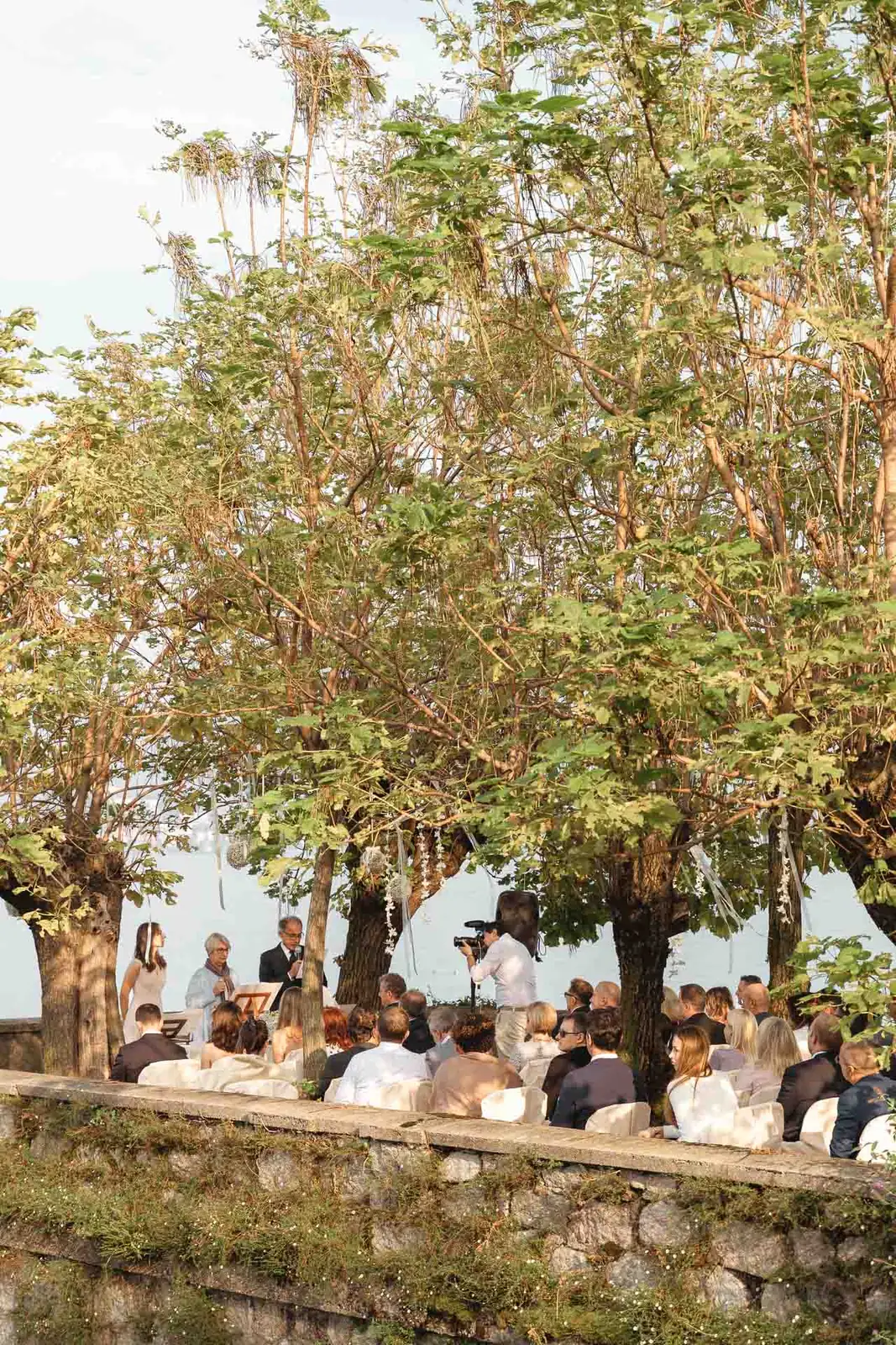Gli invitati seduti sotto gli alberi, sopra il muro in pietra in riva al lago
