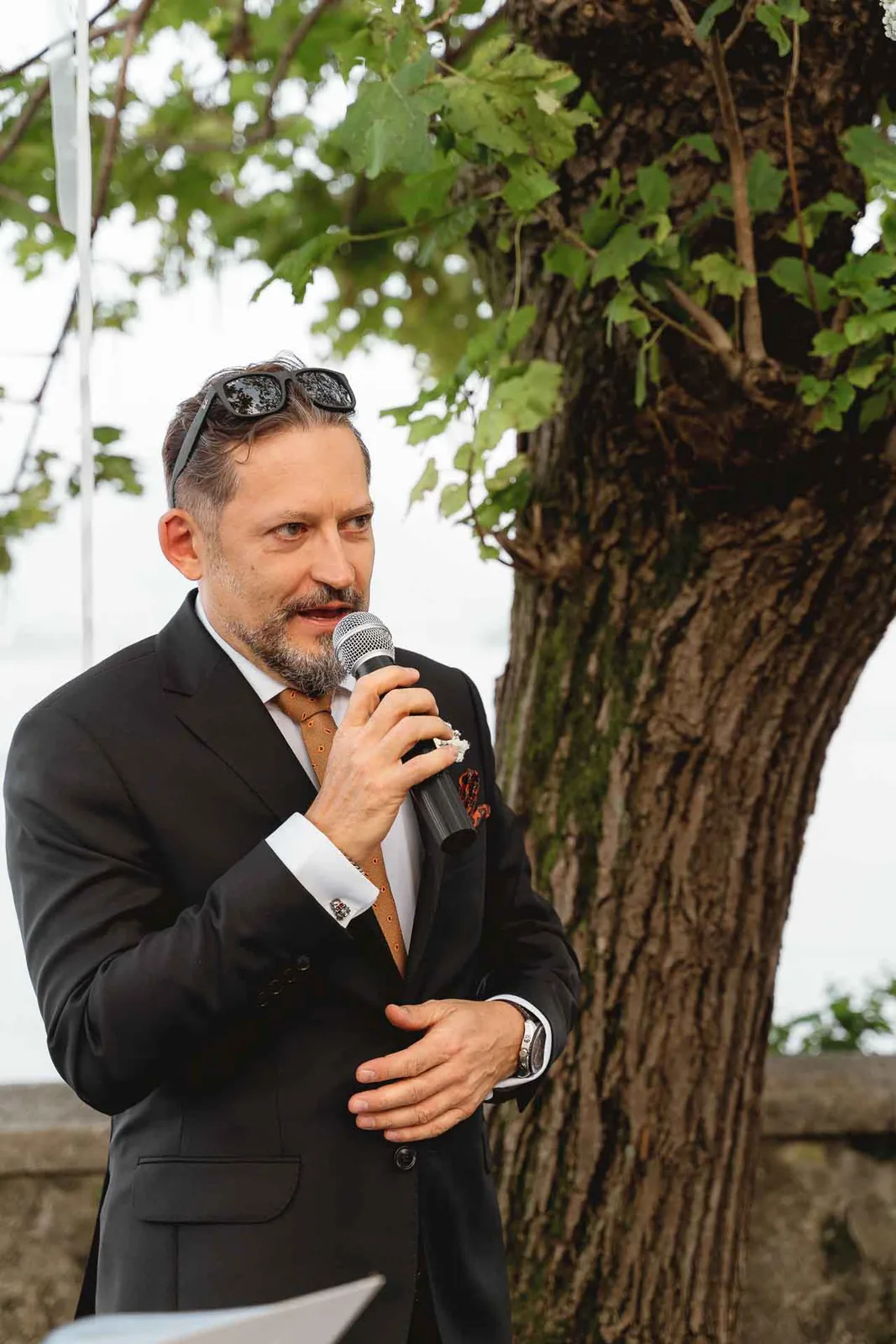 Un amico parla al microfono sotto i rami, con il lago dietro