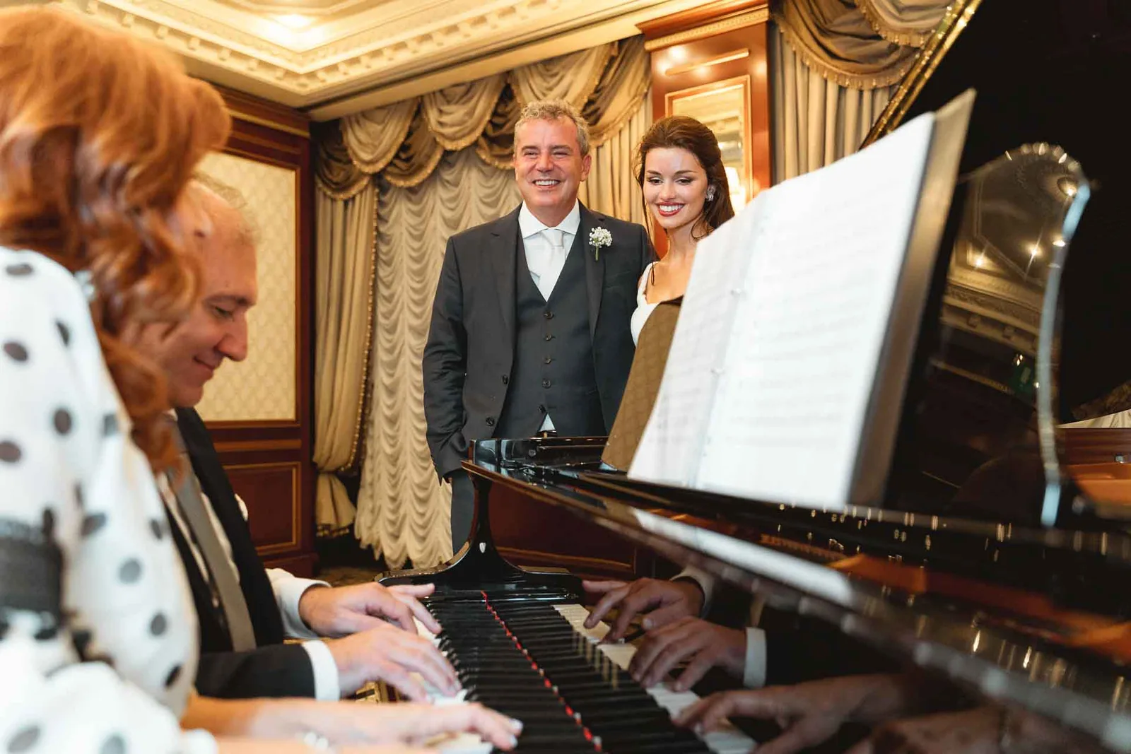 Gli sposi sorridono dietro al pianoforte, mentre una donna con l'abito a pois suona
