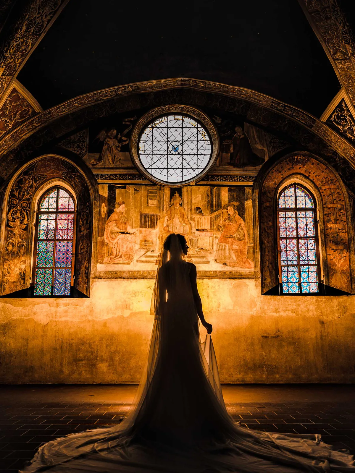 La sposa in controluce sotto una volta affrescata a San Galgano, con il velo lungo sul pavimento, il rosone e le vetrate colorate alle spalle