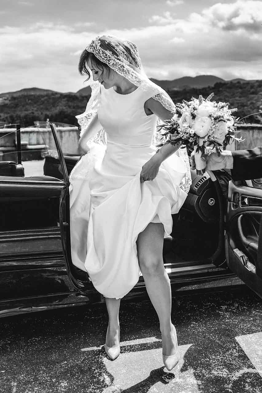 In bianco e nero, la sposa scende da un'auto scoperta tenendo l'abito, con il velo di pizzo e il bouquet