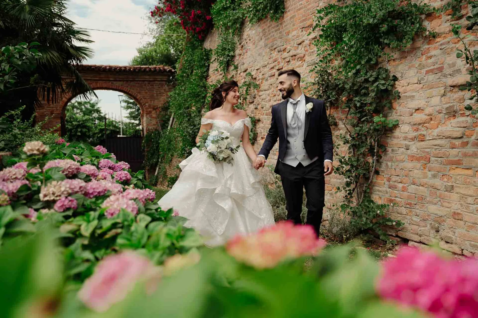 Gli sposi camminano mano nella mano tra le ortensie, lungo un muro di mattoni coperto d'edera