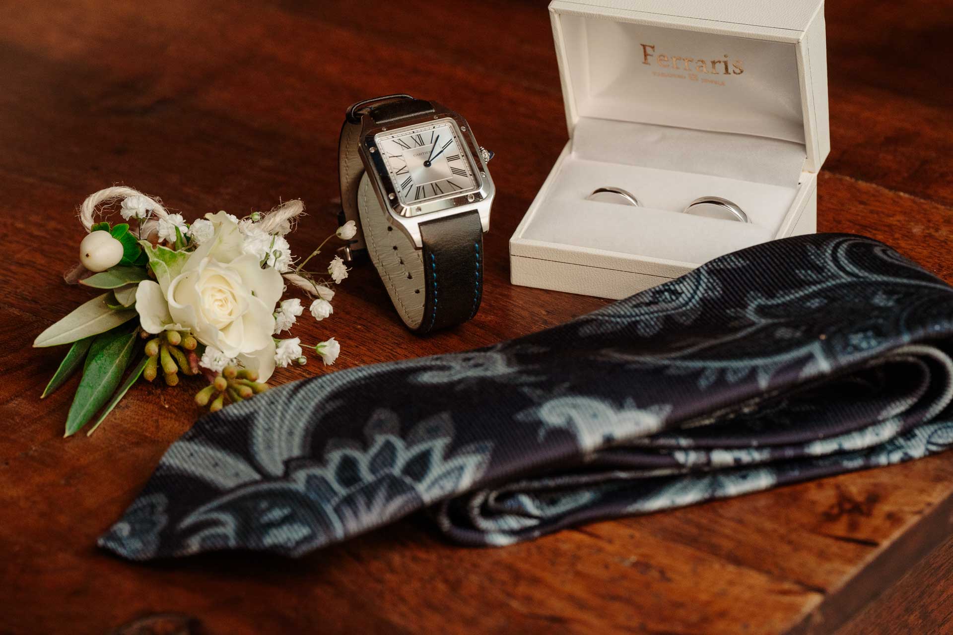 L'orologio, le fedi nella scatola, la cravatta blu a motivi paisley e il fiore all'occhiello su un tavolo di legno