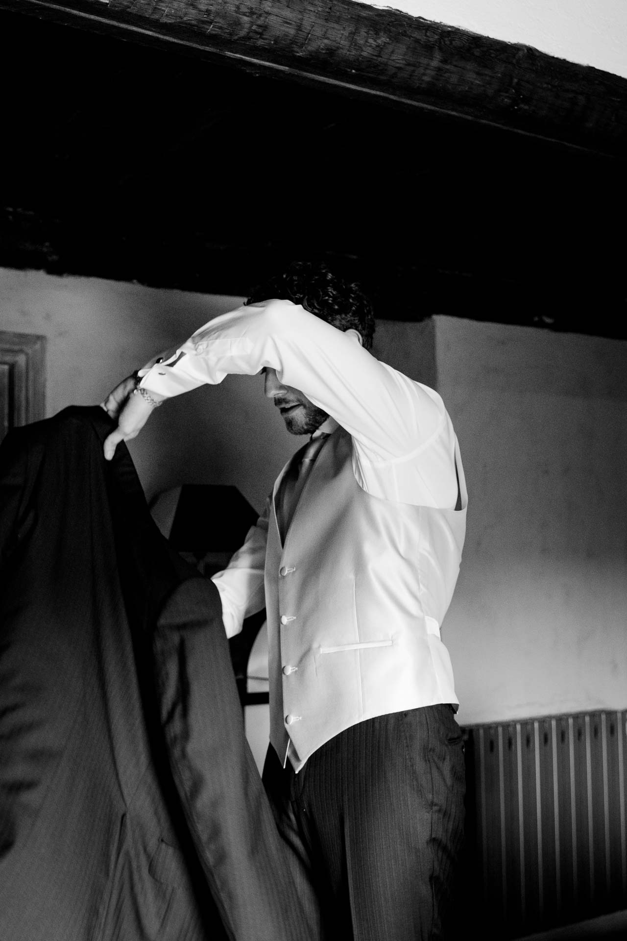 In bianco e nero, lo sposo infila la camicia sotto le travi della camera