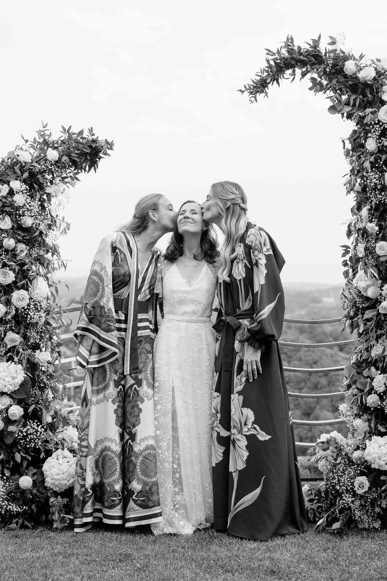 In bianco e nero, due amiche in vestaglia baciano la sposa sulle guance, sotto l'arco di fiori