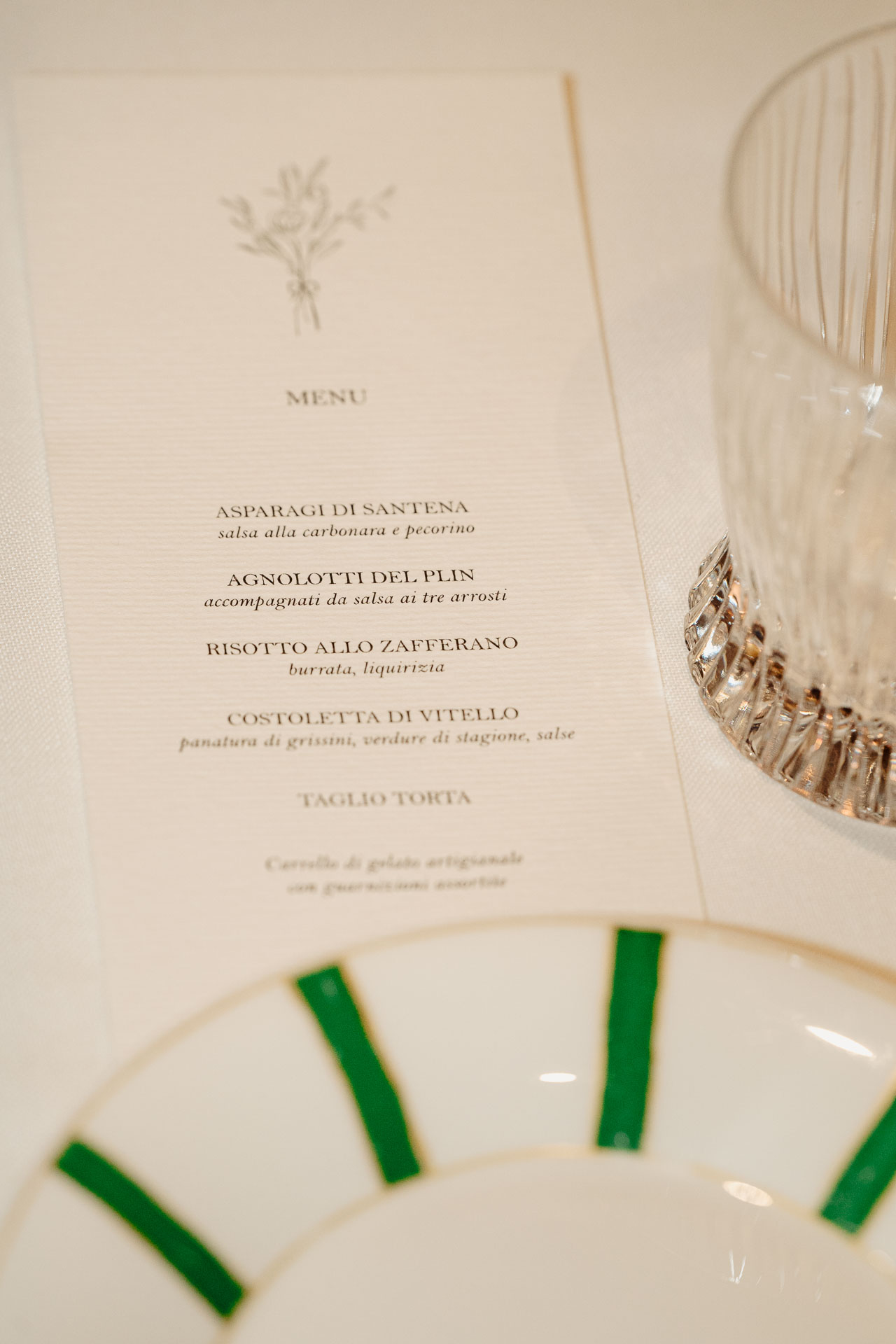 Il menu della cena, tra il piatto a raggi verdi e un bicchiere di cristallo