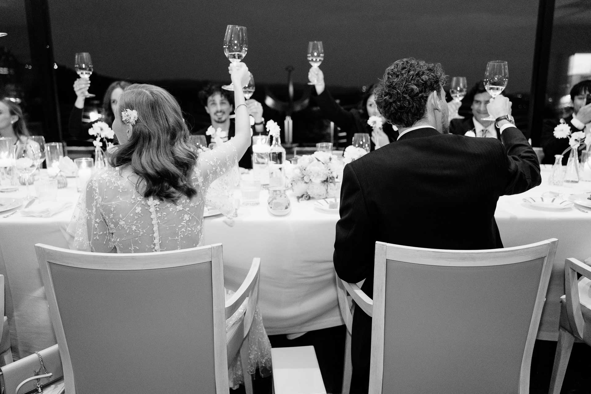 In bianco e nero, gli invitati alzano i calici per il brindisi, alle spalle degli sposi