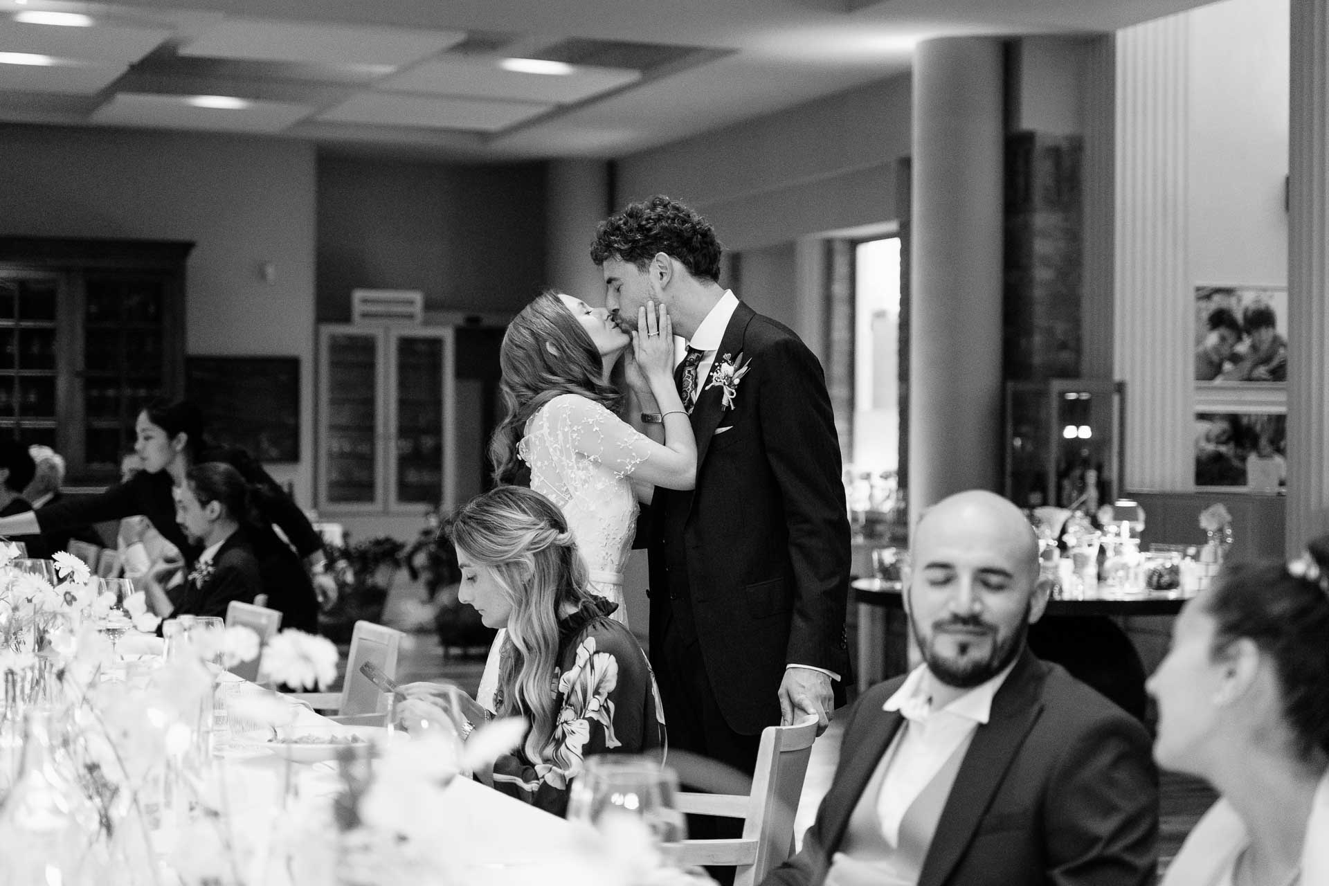 In bianco e nero, gli sposi si baciano in piedi alla tavola, tra gli invitati