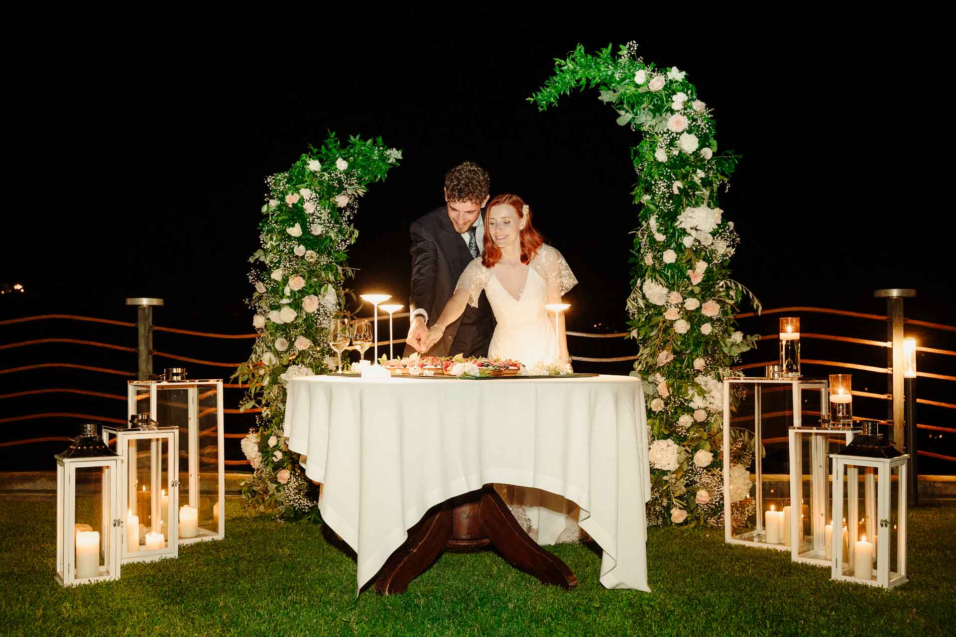 Gli sposi tagliano la torta sul prato, di notte, sotto l'arco di fiori e tra le lanterne