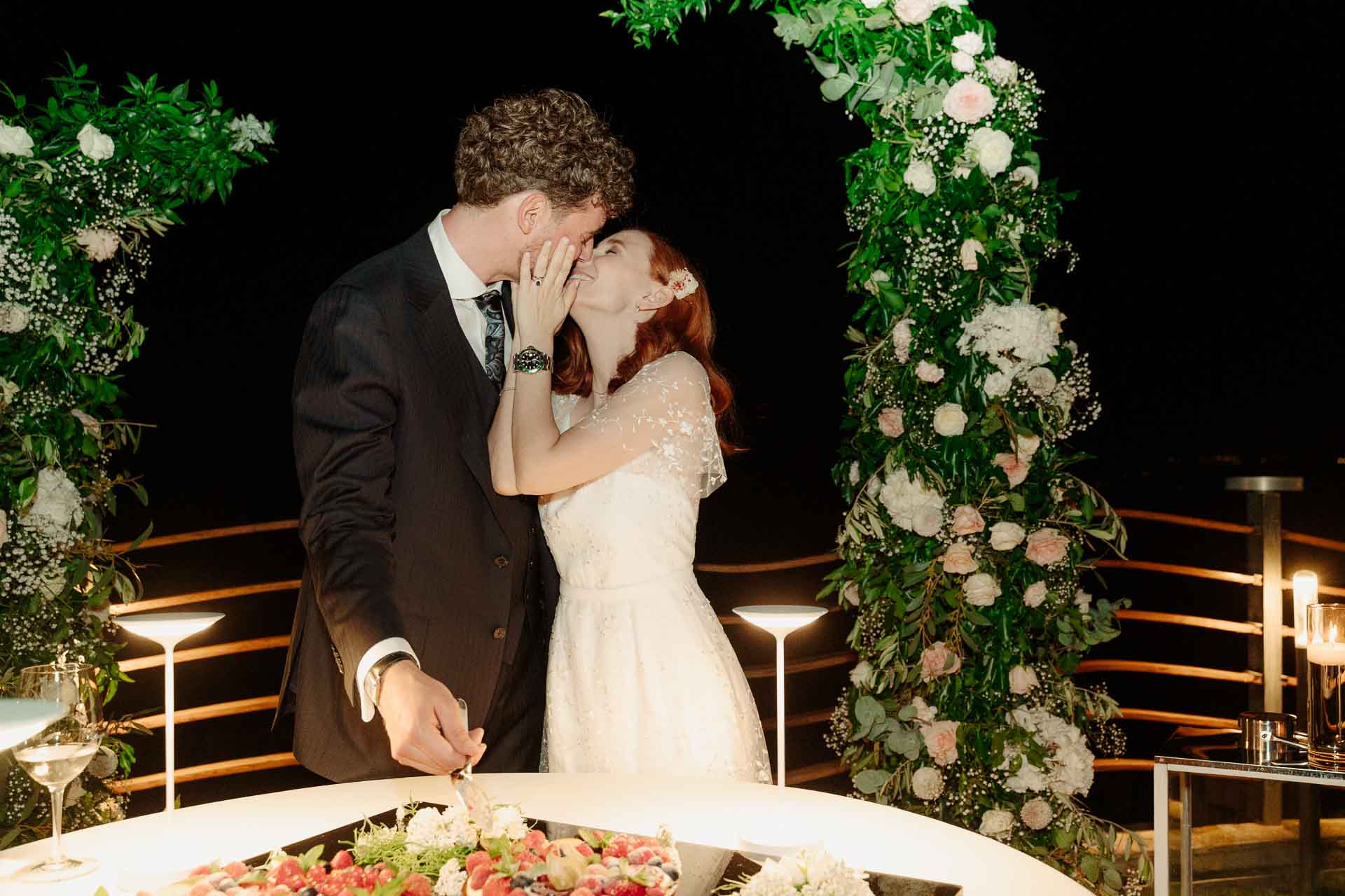 Gli sposi si baciano mentre tagliano la crostata, sotto l'arco di fiori