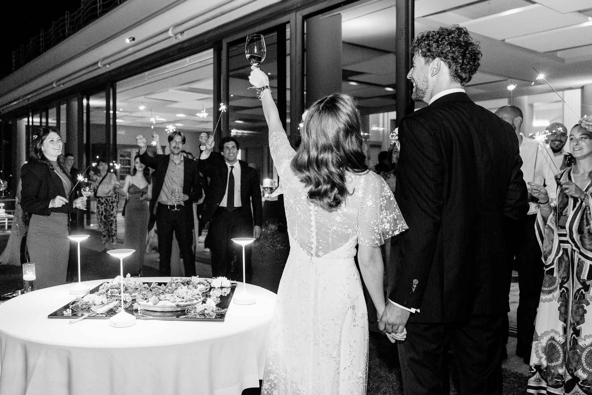 In bianco e nero, la sposa alza il calice verso gli invitati, mano nella mano con lo sposo