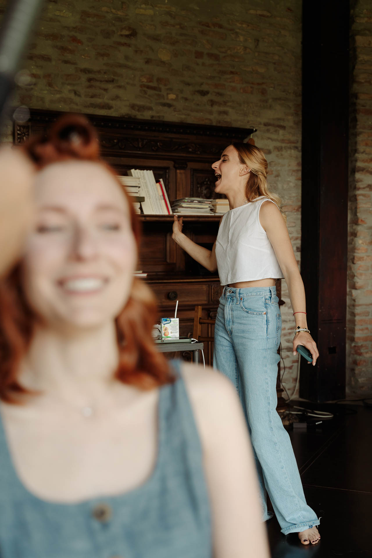 La sposa sorride in primo piano, mentre un'amica in jeans ride accanto alla libreria