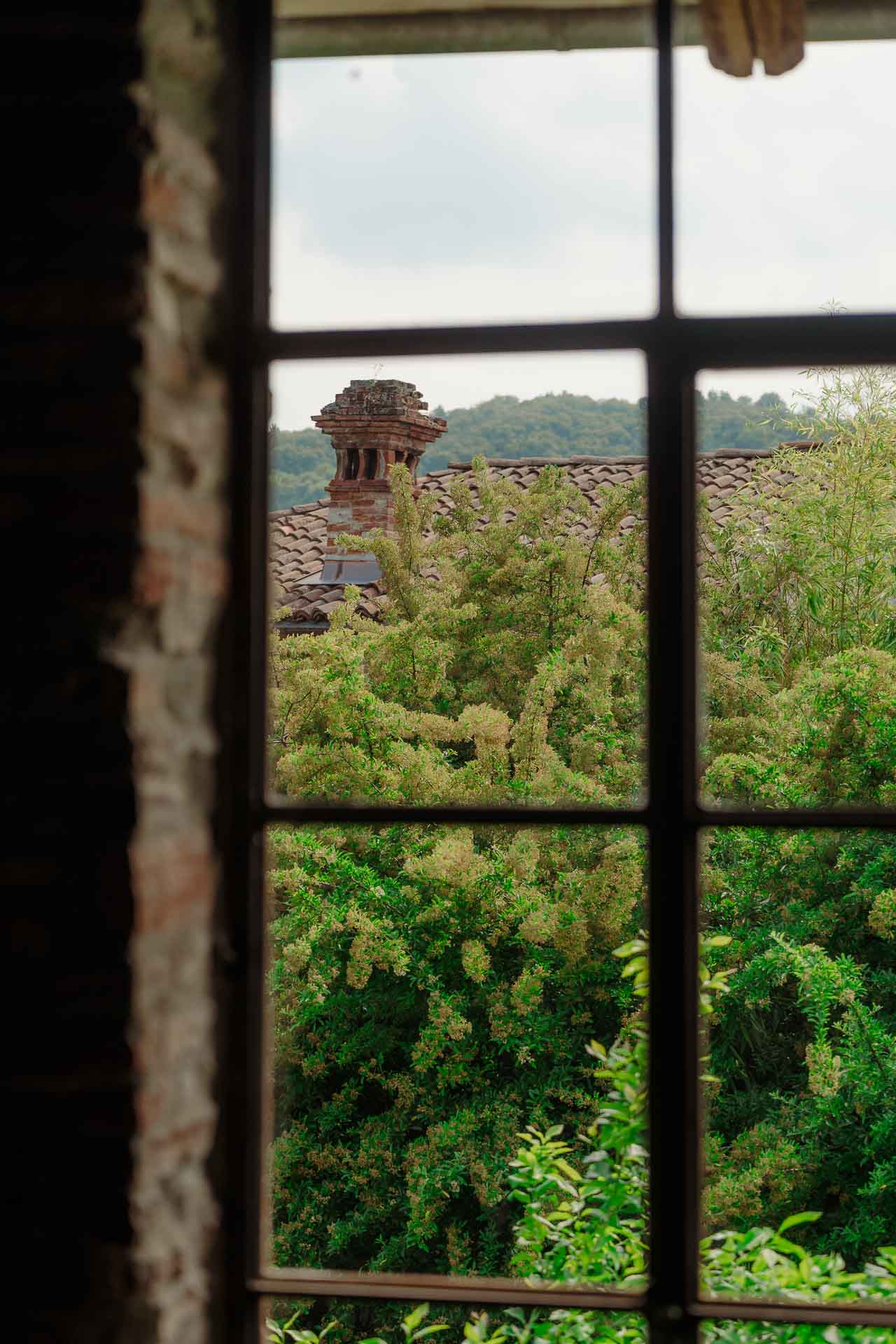 Dalla finestra, gli alberi e un vecchio comignolo sui tetti