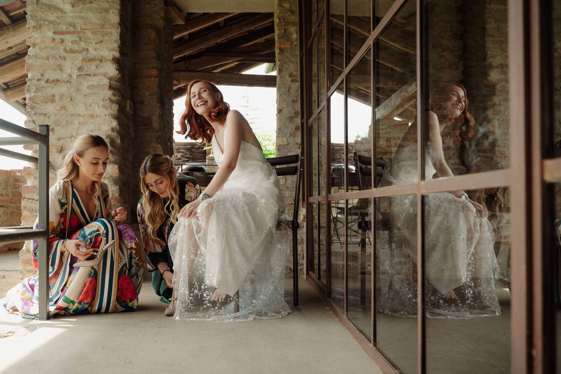 La sposa seduta mentre le amiche le allacciano le scarpe, riflesse nella vetrata