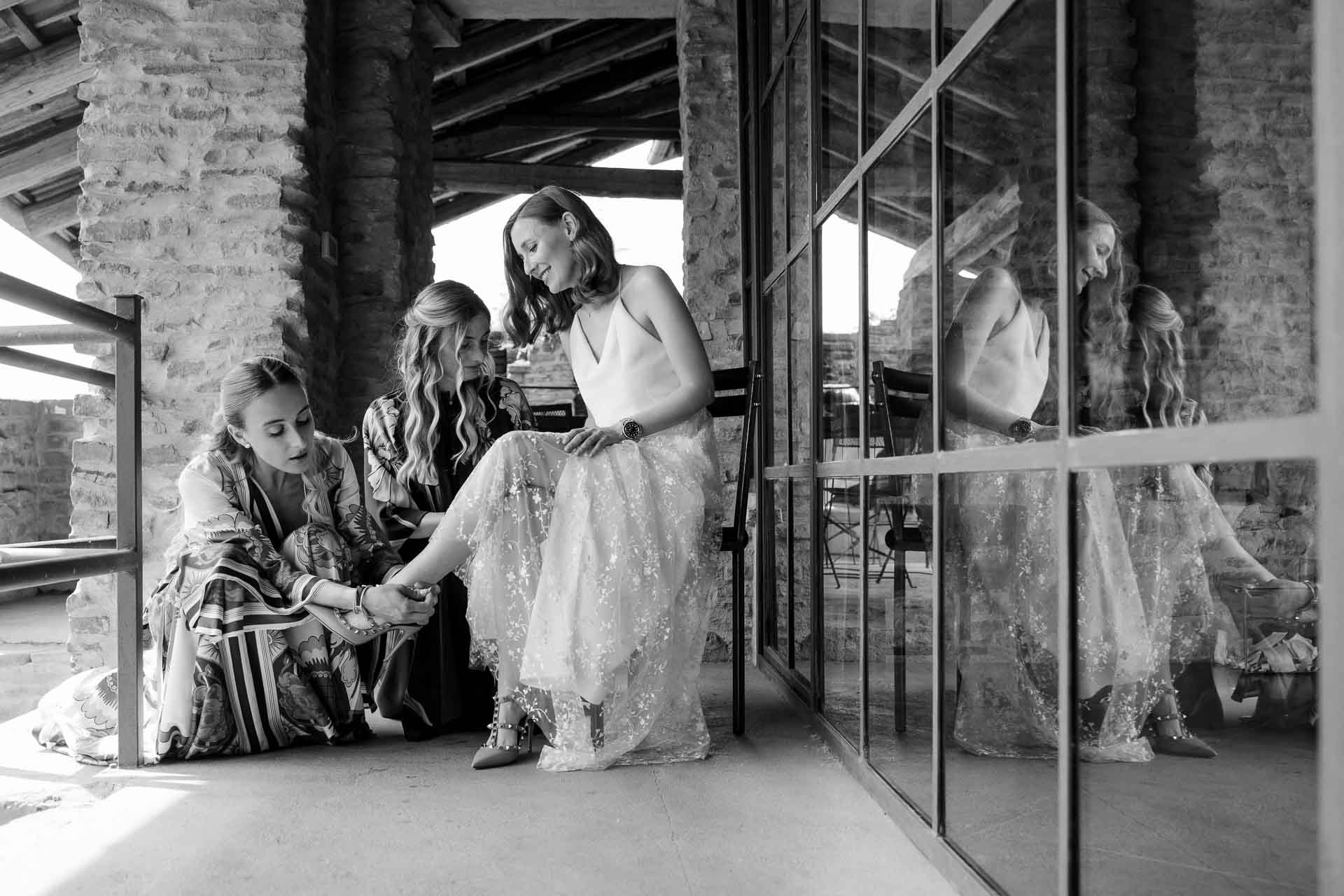 In bianco e nero, le amiche allacciano le scarpe alla sposa, riflesse nella vetrata