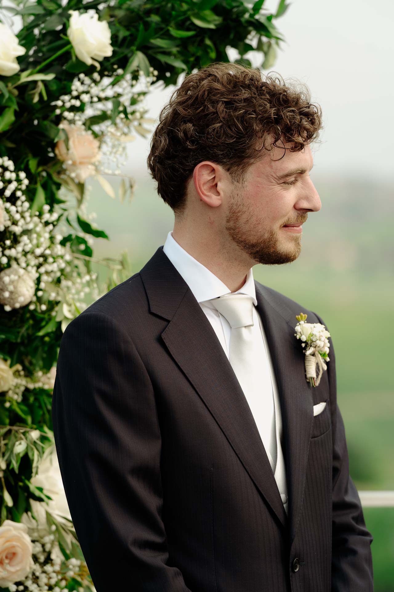 Lo sposo ascolta a sguardo basso, davanti all'arco di fiori