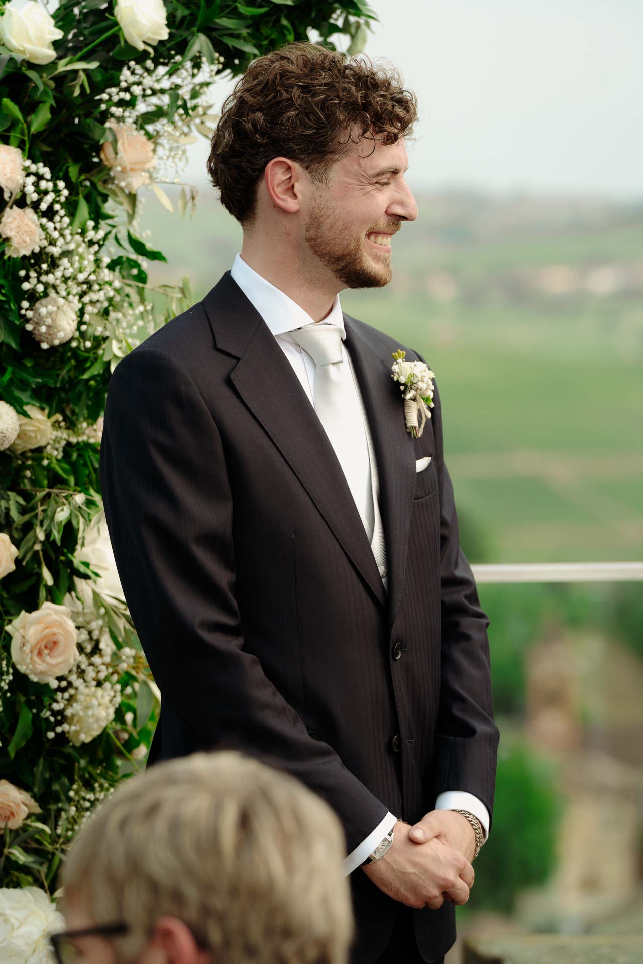 Lo sposo sorride accanto all'arco di rose, con le colline alle spalle