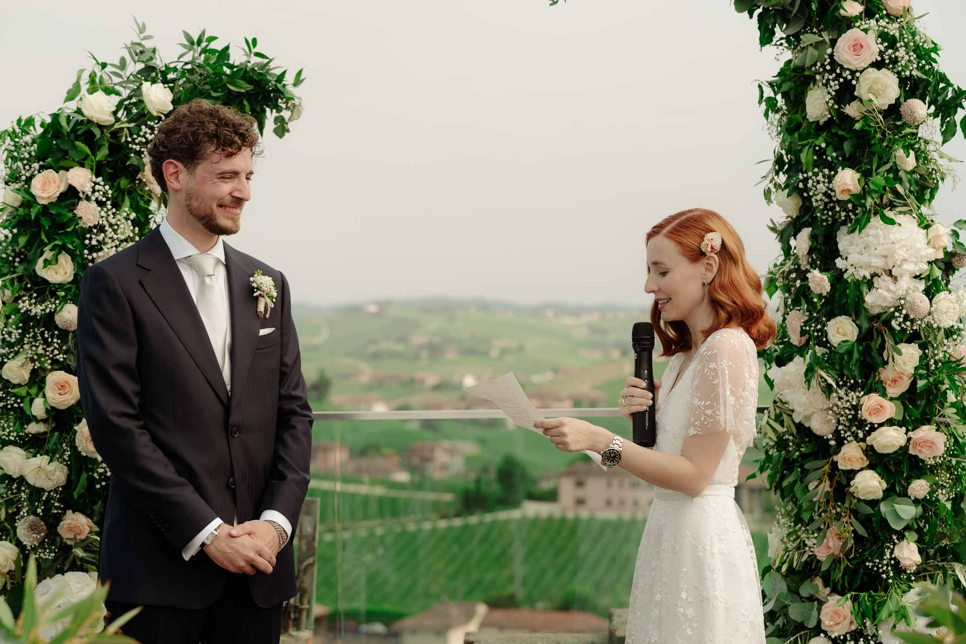 La sposa legge le promesse allo sposo tra i due archi di fiori, sopra le vigne
