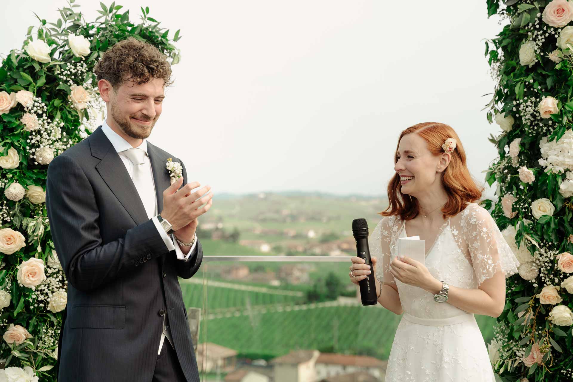 Gli sposi ridono tra i due archi di fiori, sopra le vigne