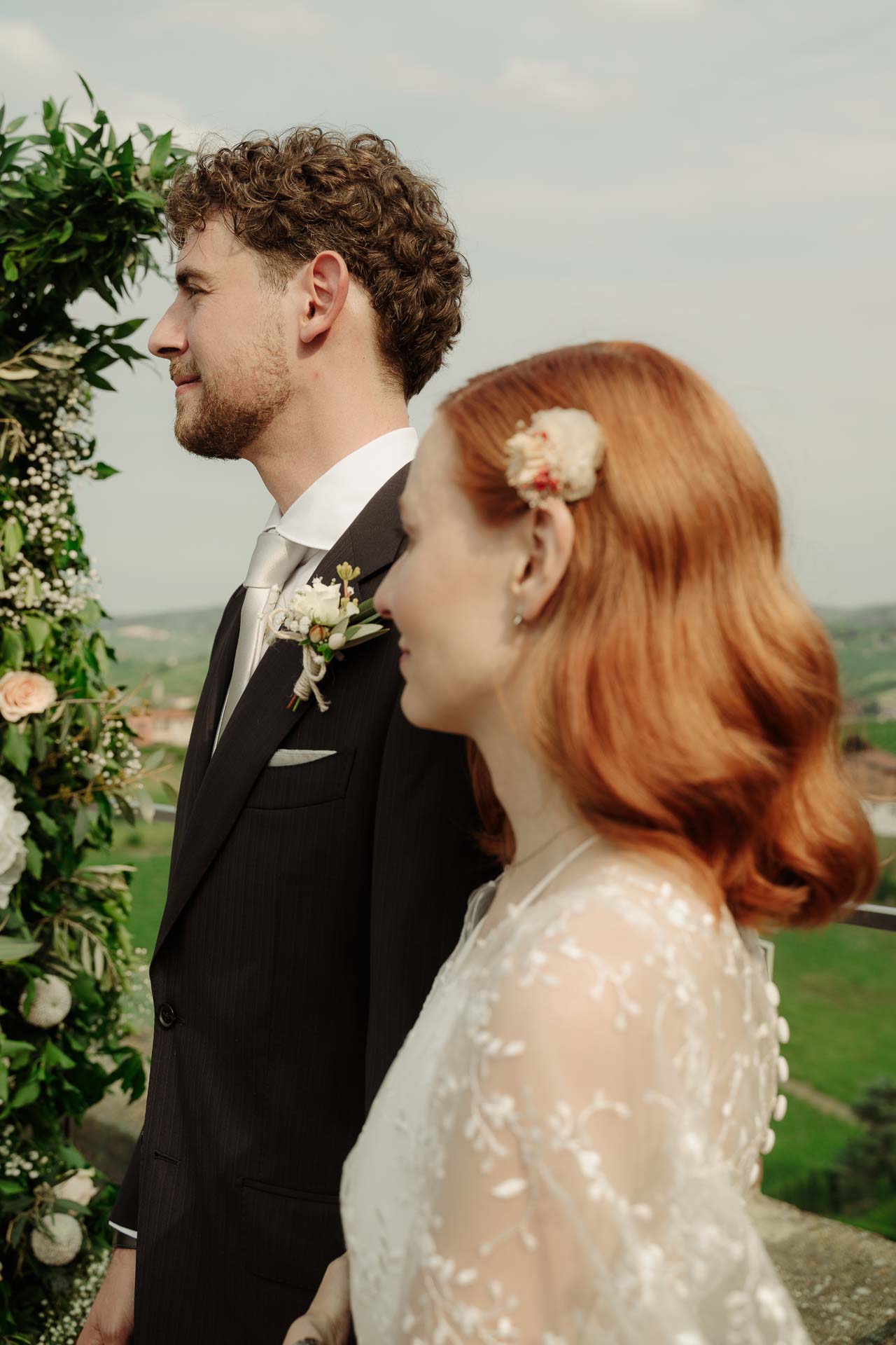 Gli sposi di profilo, lui guarda lontano, lei sorride con un fiore tra i capelli
