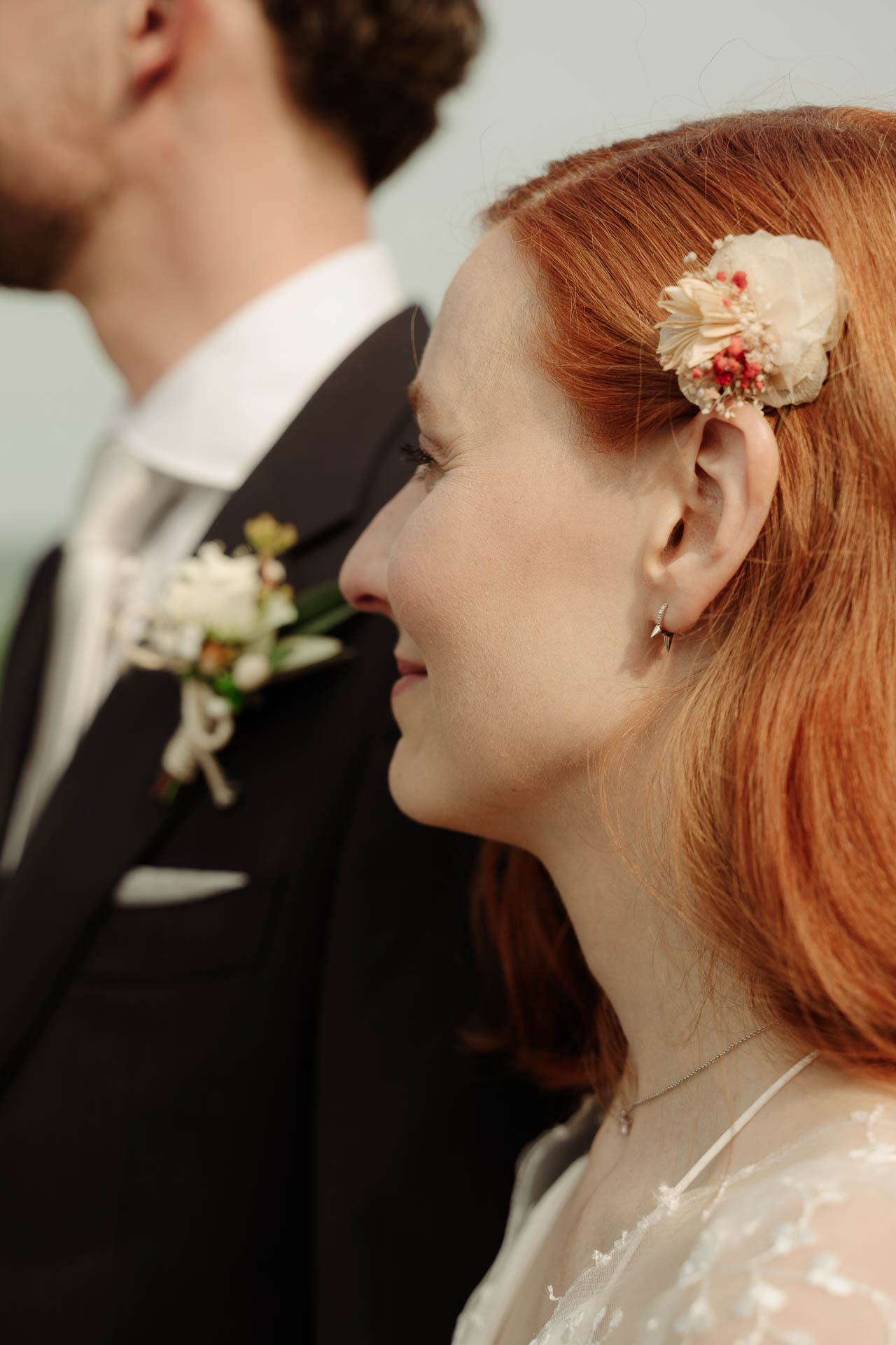 La sposa sorride di profilo accanto allo sposo, con il fiore tra i capelli rossi