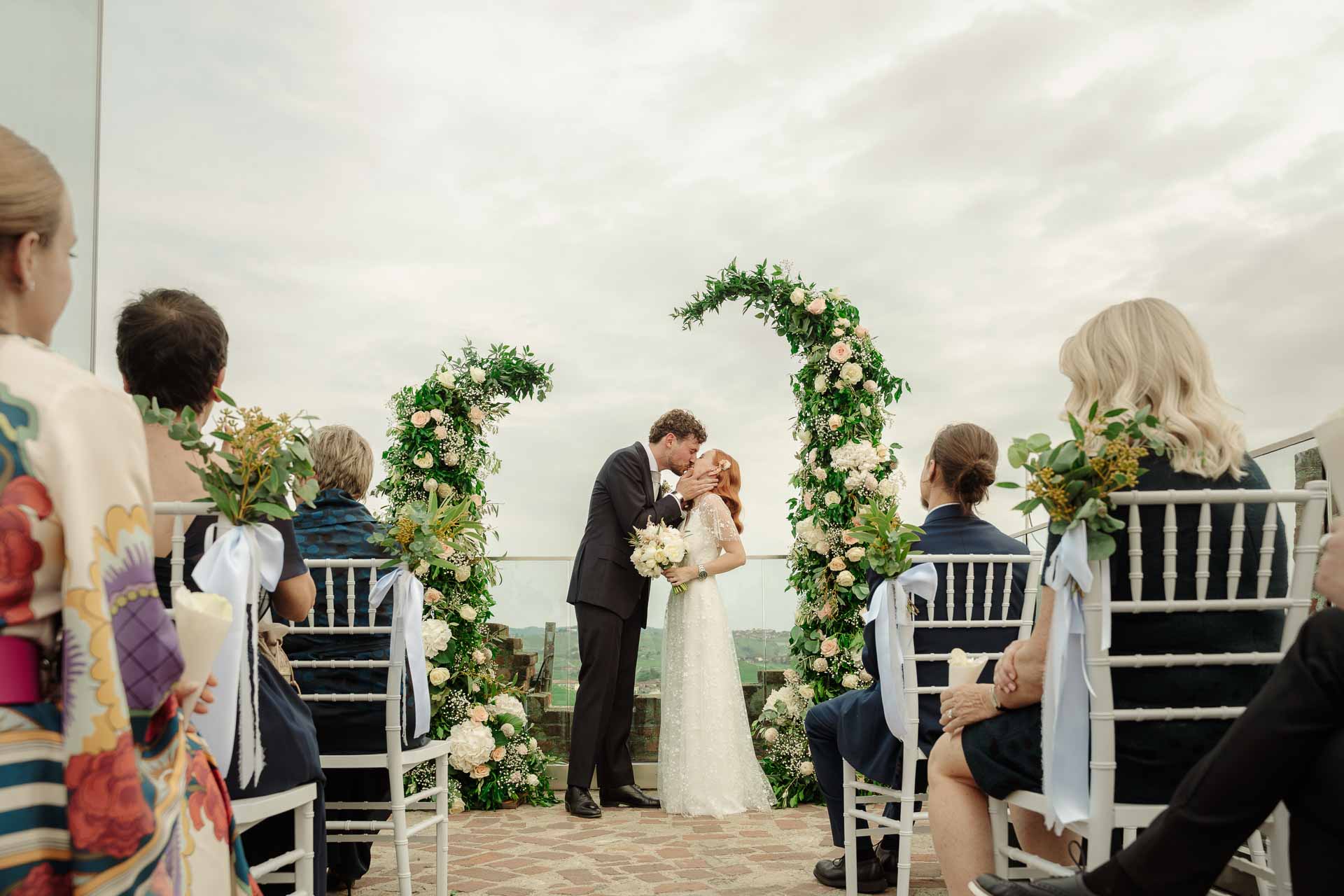 Gli sposi si baciano tra i due archi di fiori, visti dal fondo tra gli invitati