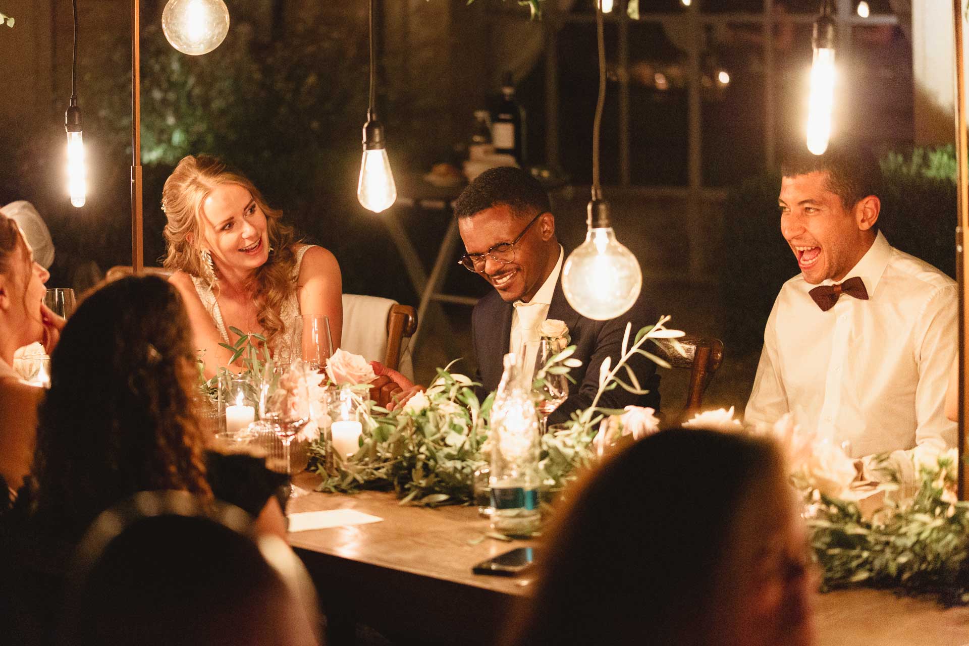 Gli sposi e gli amici ridono a tavola, sotto le lampadine