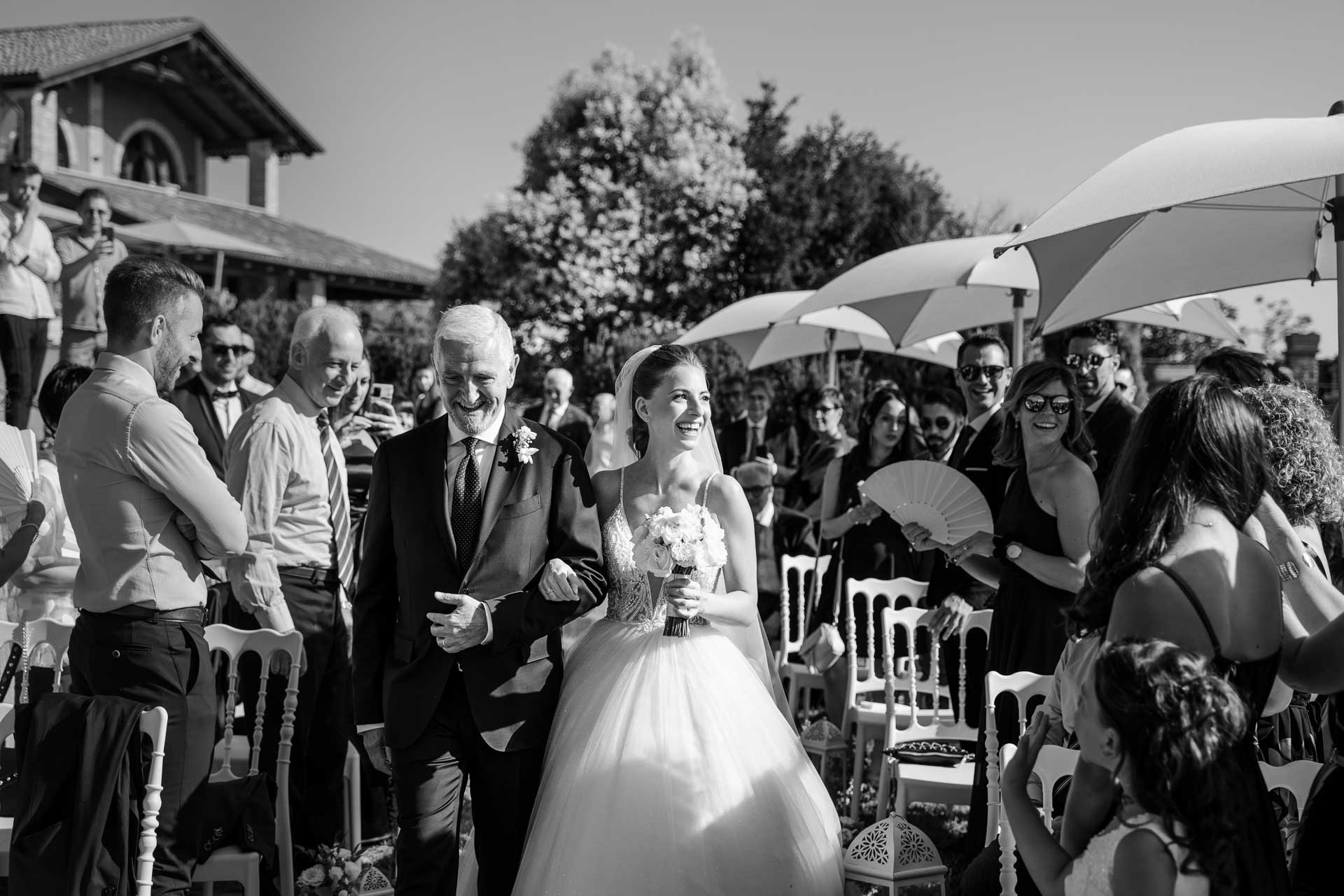 In bianco e nero, la sposa sorride al braccio del papà, tra i ventagli degli invitati