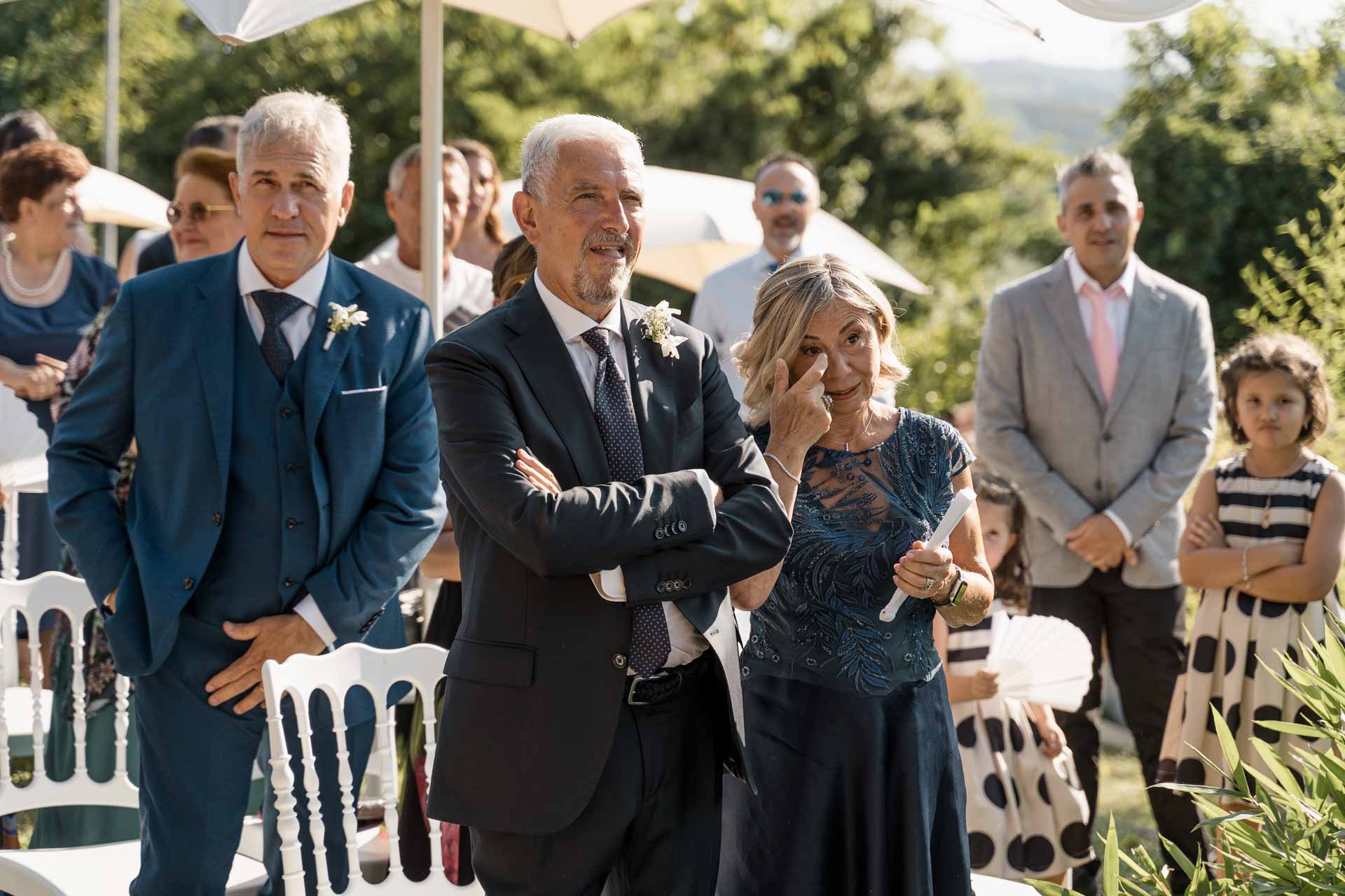 I genitori in piedi tra gli invitati, e la mamma si asciuga una lacrima