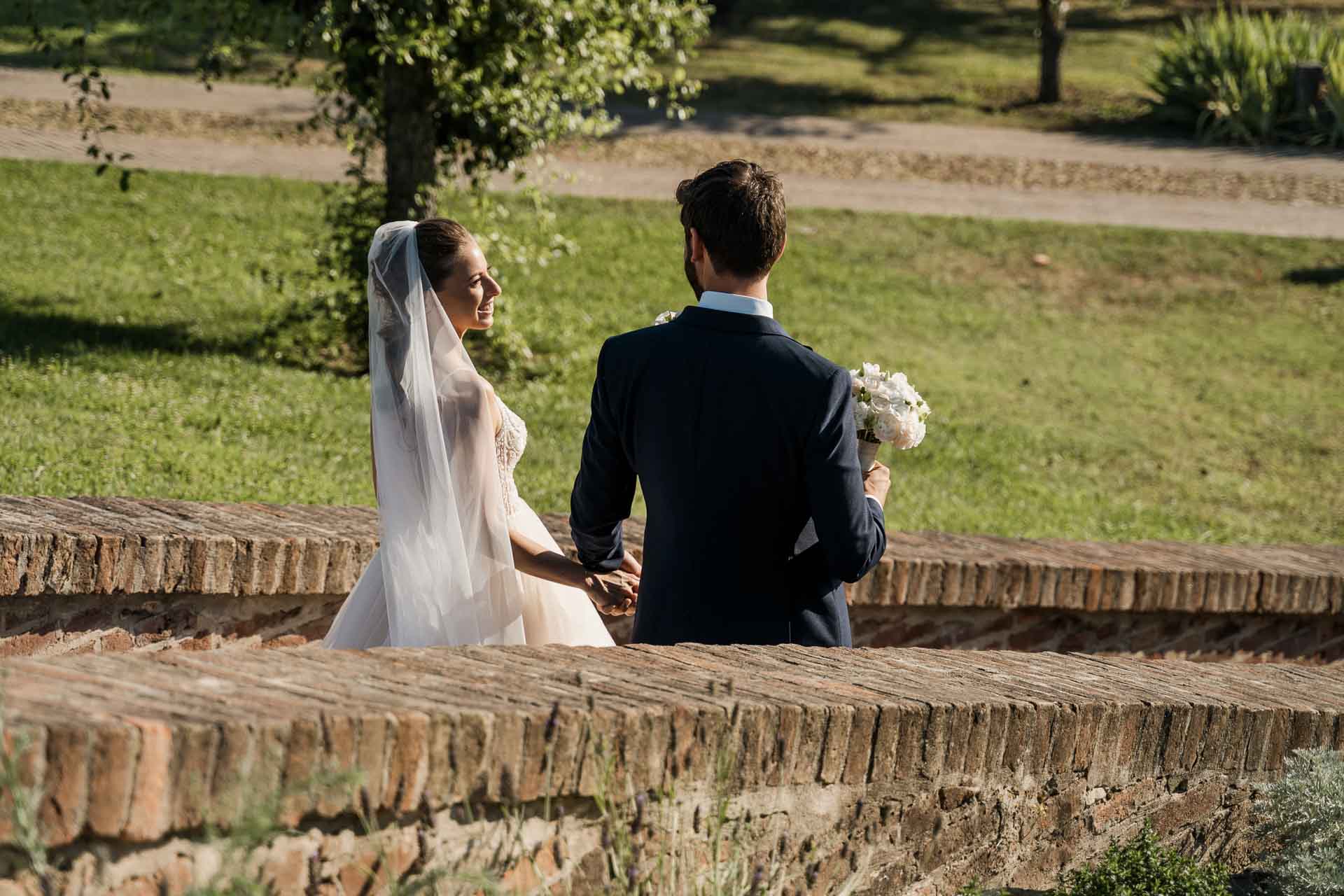 Gli sposi di spalle sul muretto di mattoni, verso il prato
