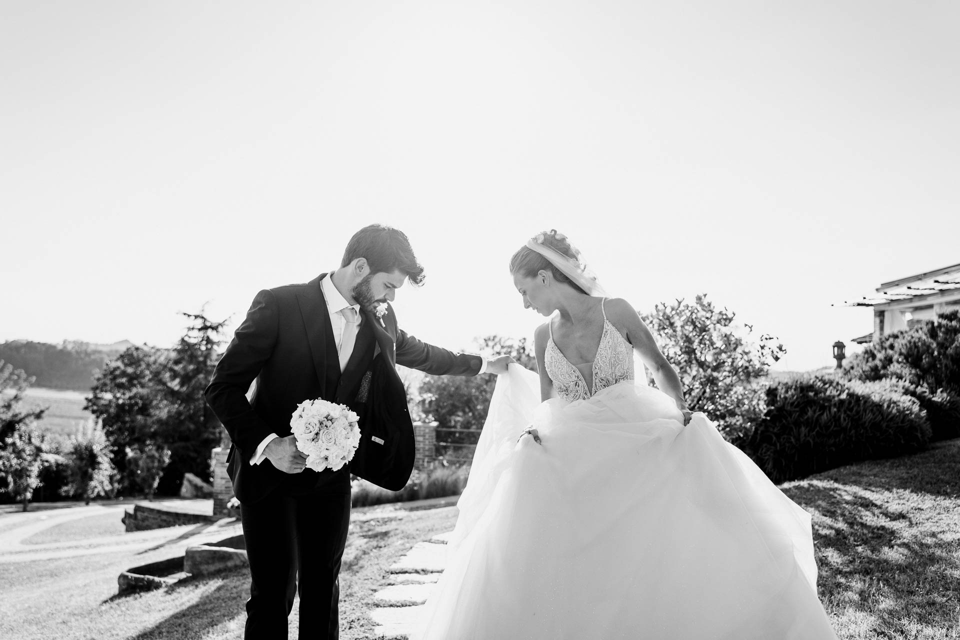 In bianco e nero, lo sposo aiuta la sposa con l'abito, nel prato