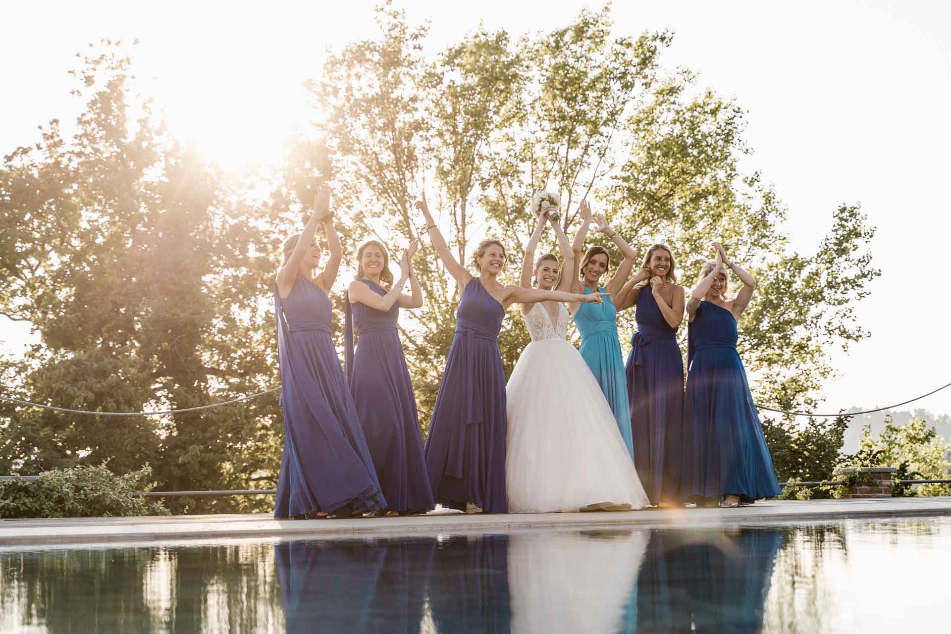 La sposa e le amiche in blu e azzurro a braccia alzate sul bordo della piscina