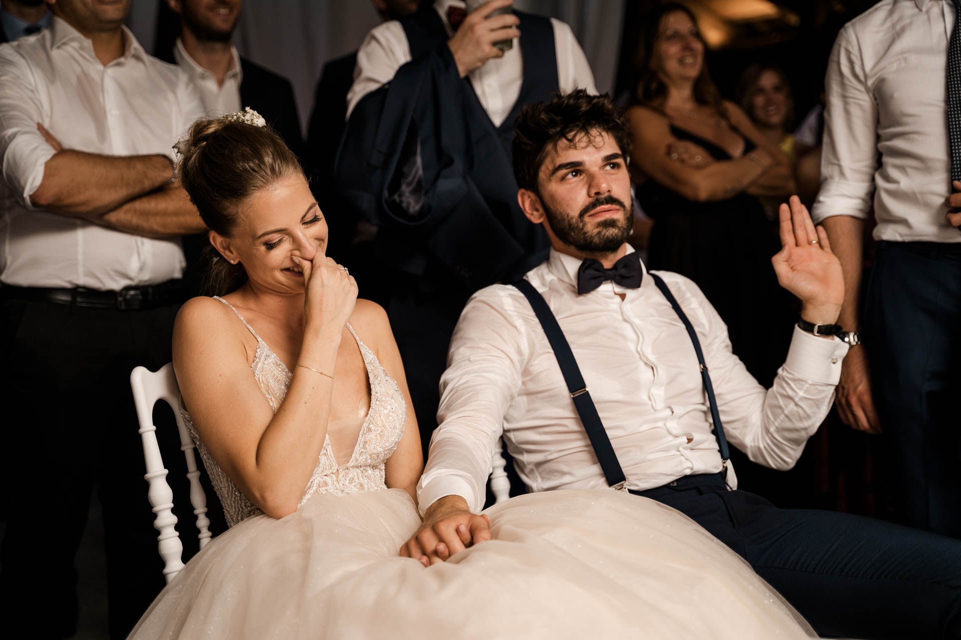 Gli sposi seduti in mezzo agli invitati: la sposa ride, lo sposo alza la mano