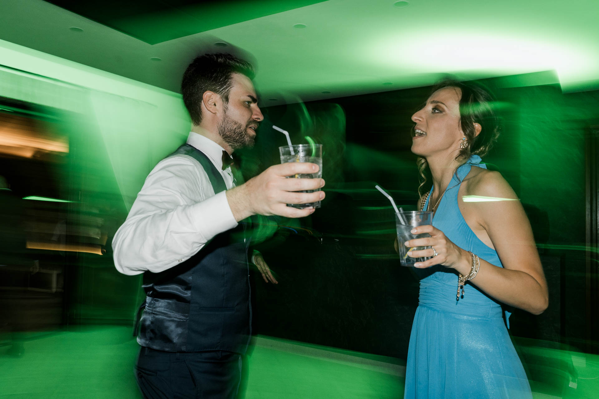 Lo sposo e l'amica in azzurro ballano con il bicchiere in mano, nelle luci verdi