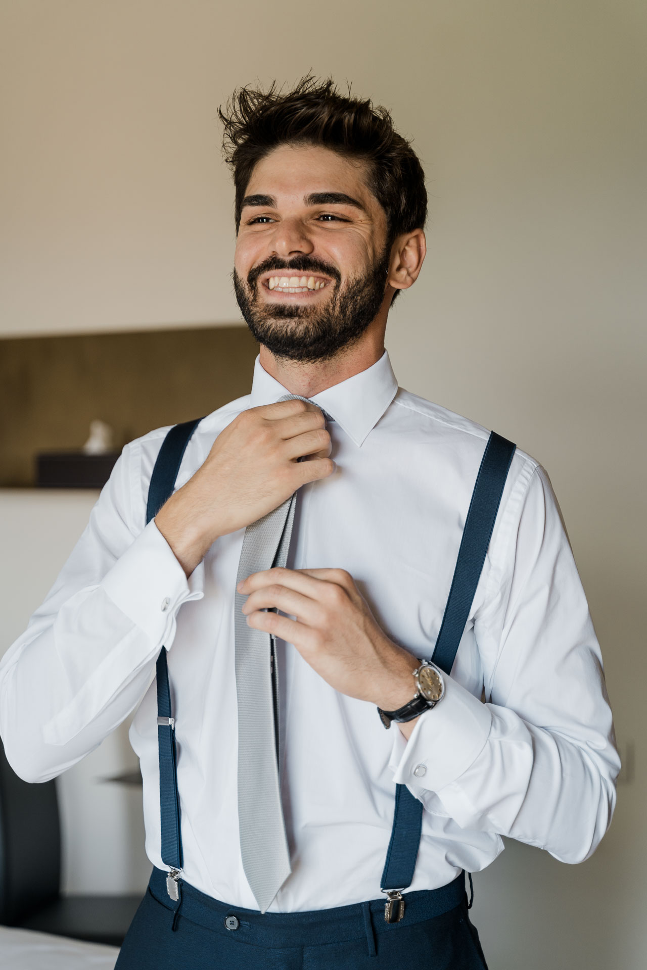 Lo sposo sorride mentre stringe il nodo della cravatta, con le bretelle blu