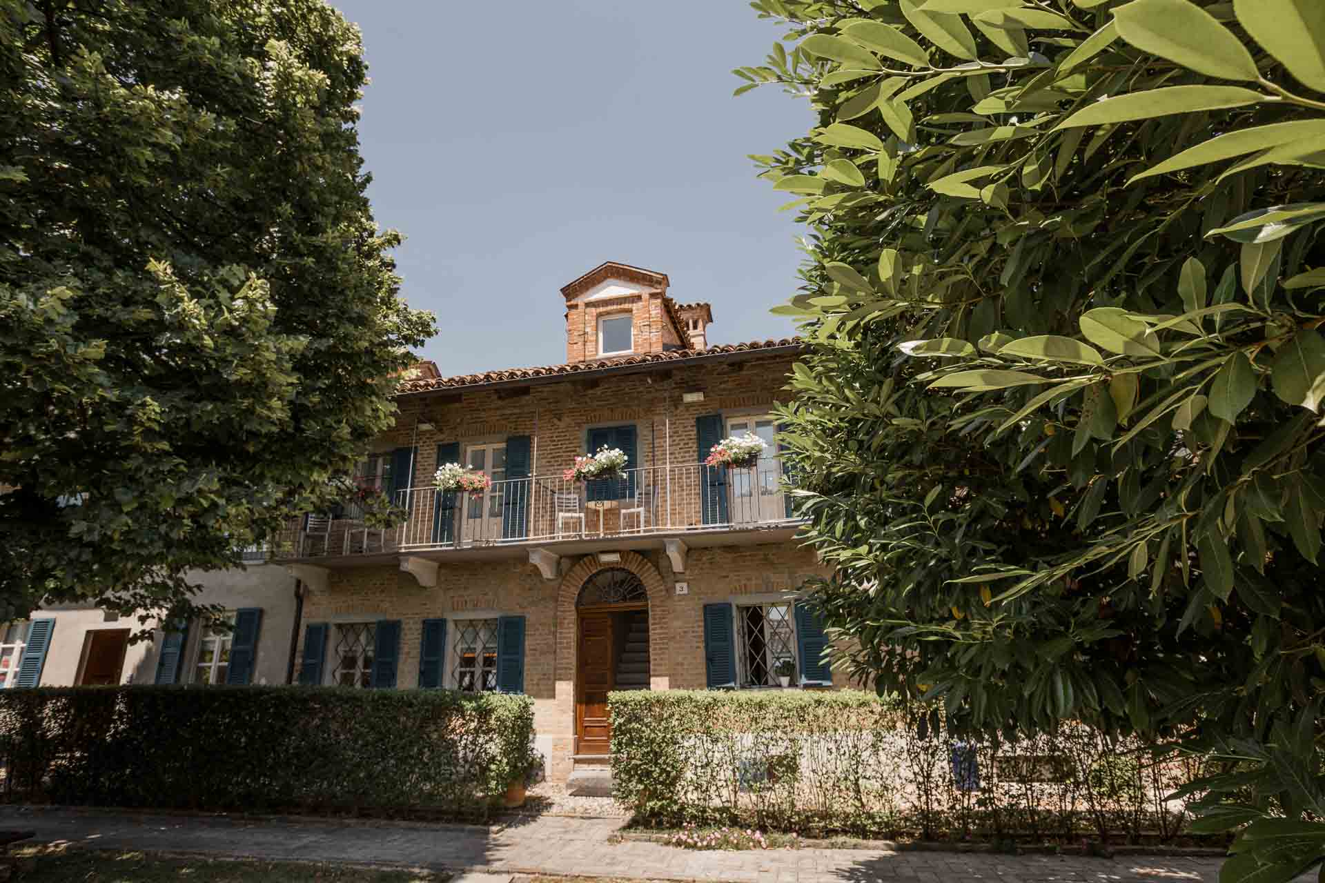 La facciata della cascina della Madernassa, a Guarene, tra gli alberi, con le persiane blu e i balconi fioriti