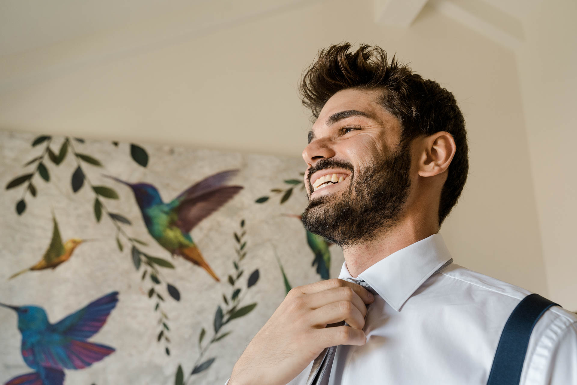Lo sposo ride sistemandosi la cravatta, tra i colibrì dipinti sul muro