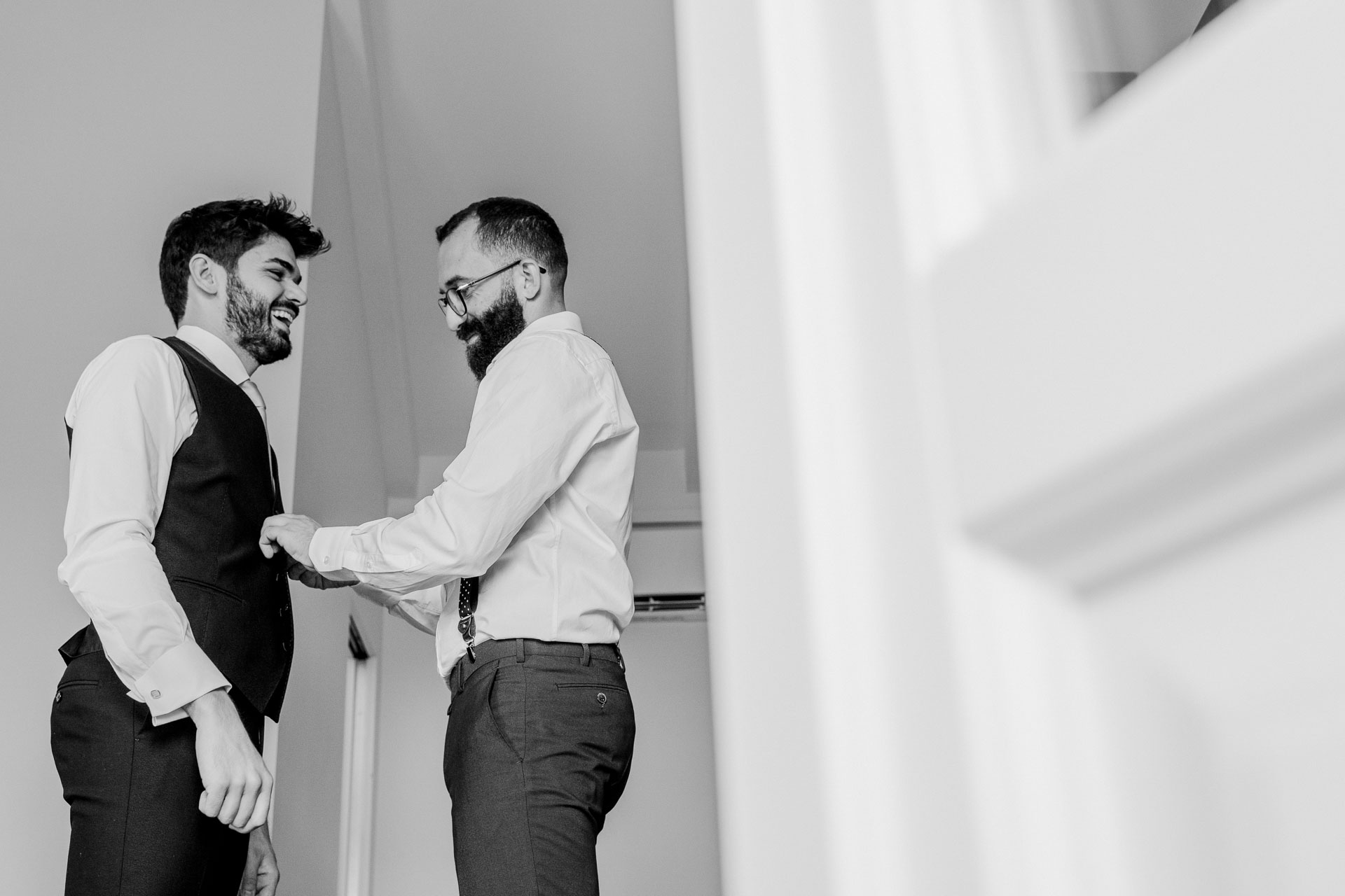 In bianco e nero, l'amico allaccia il gilet allo sposo, che ride