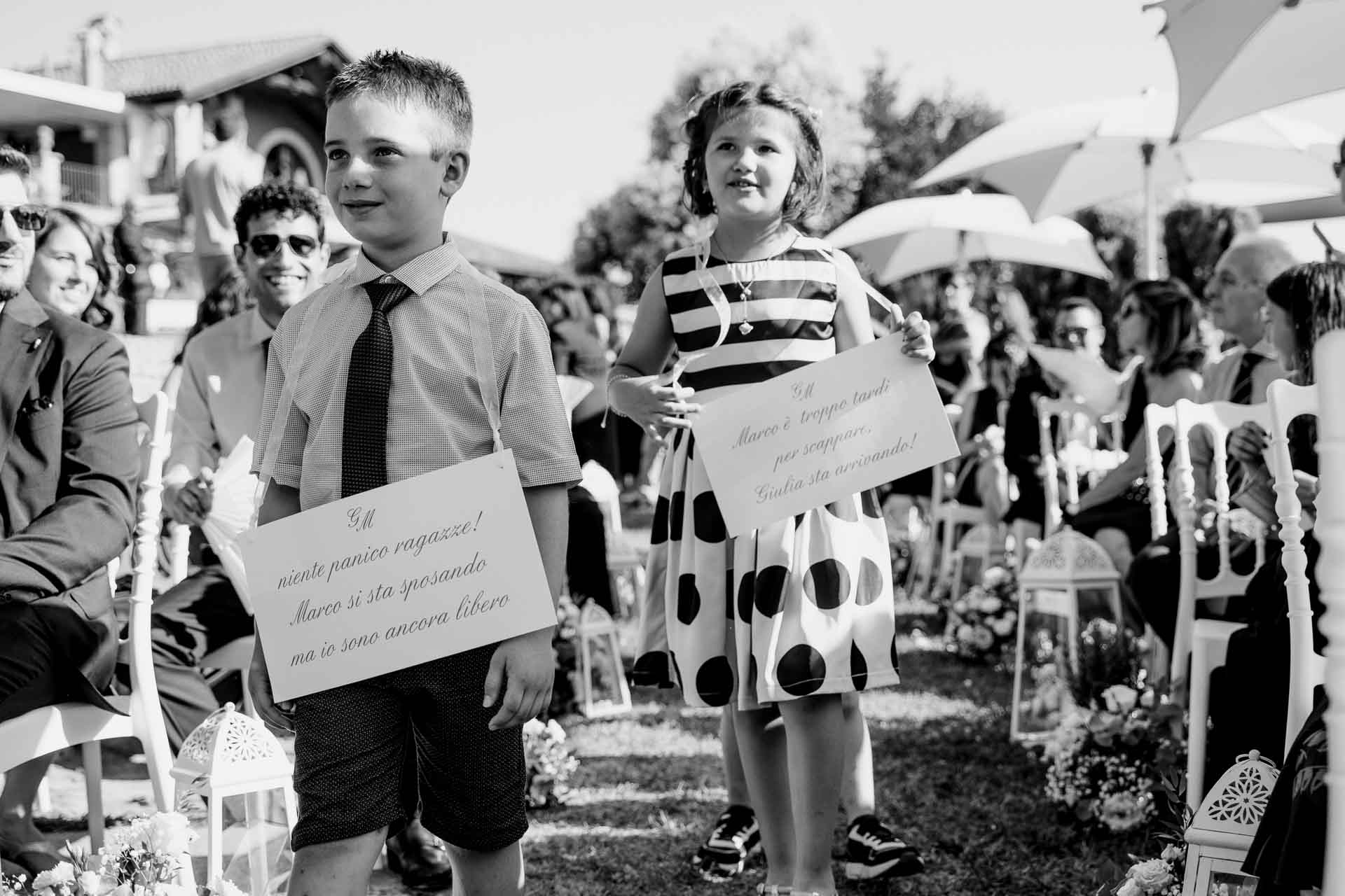 In bianco e nero, due bambini con i cartelli: "Marco è troppo tardi per scappare, Giulia sta arrivando!"