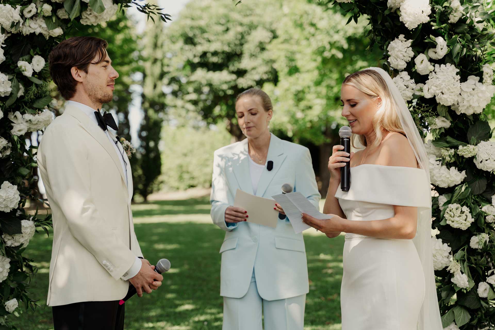 La sposa legge le promesse al microfono, sotto l'arco di fiori