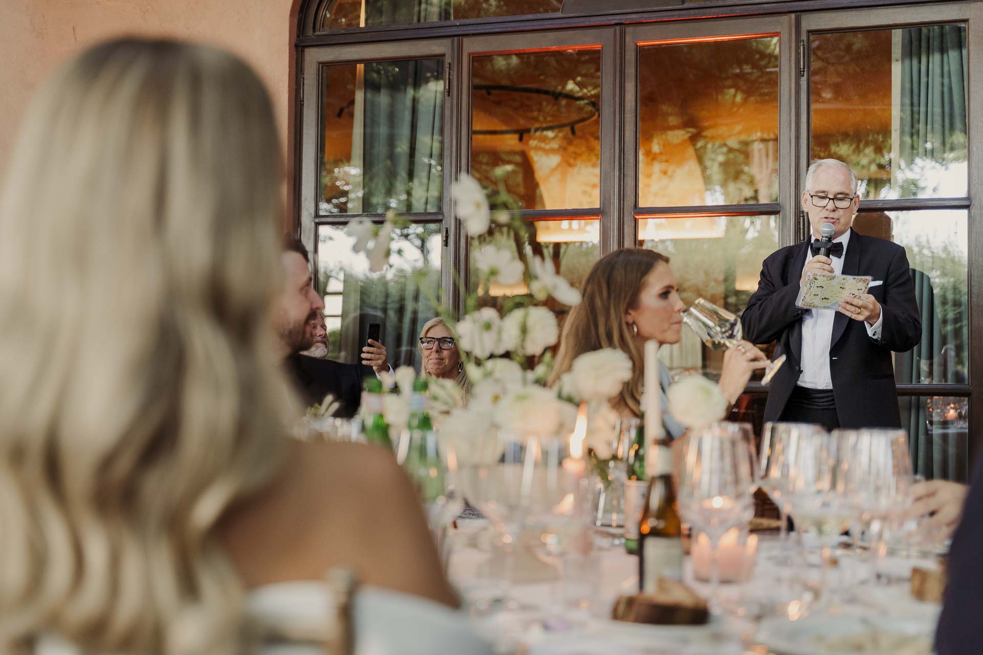 Un signore fa un discorso al microfono, visto oltre i capelli di un'invitata