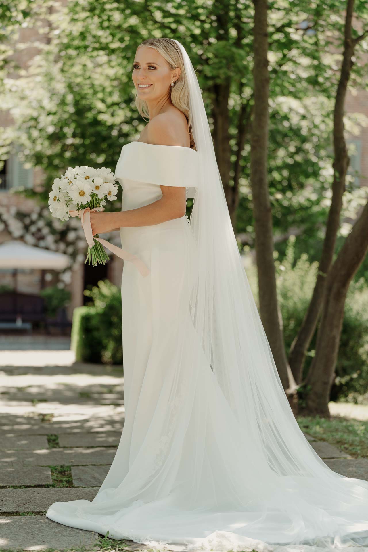 La sposa con il bouquet di margherite e il velo lungo, sotto gli alberi