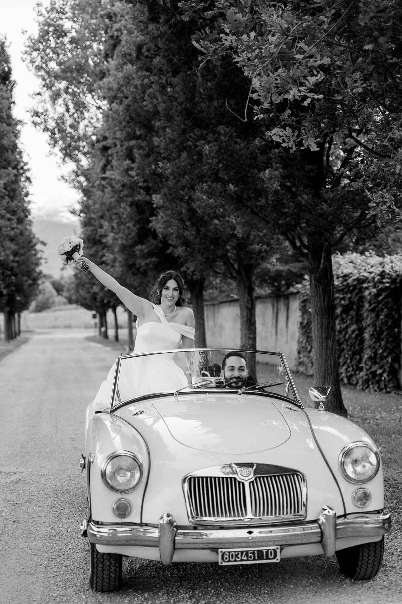 In bianco e nero, la sposa saluta in piedi sull'auto d'epoca nel viale