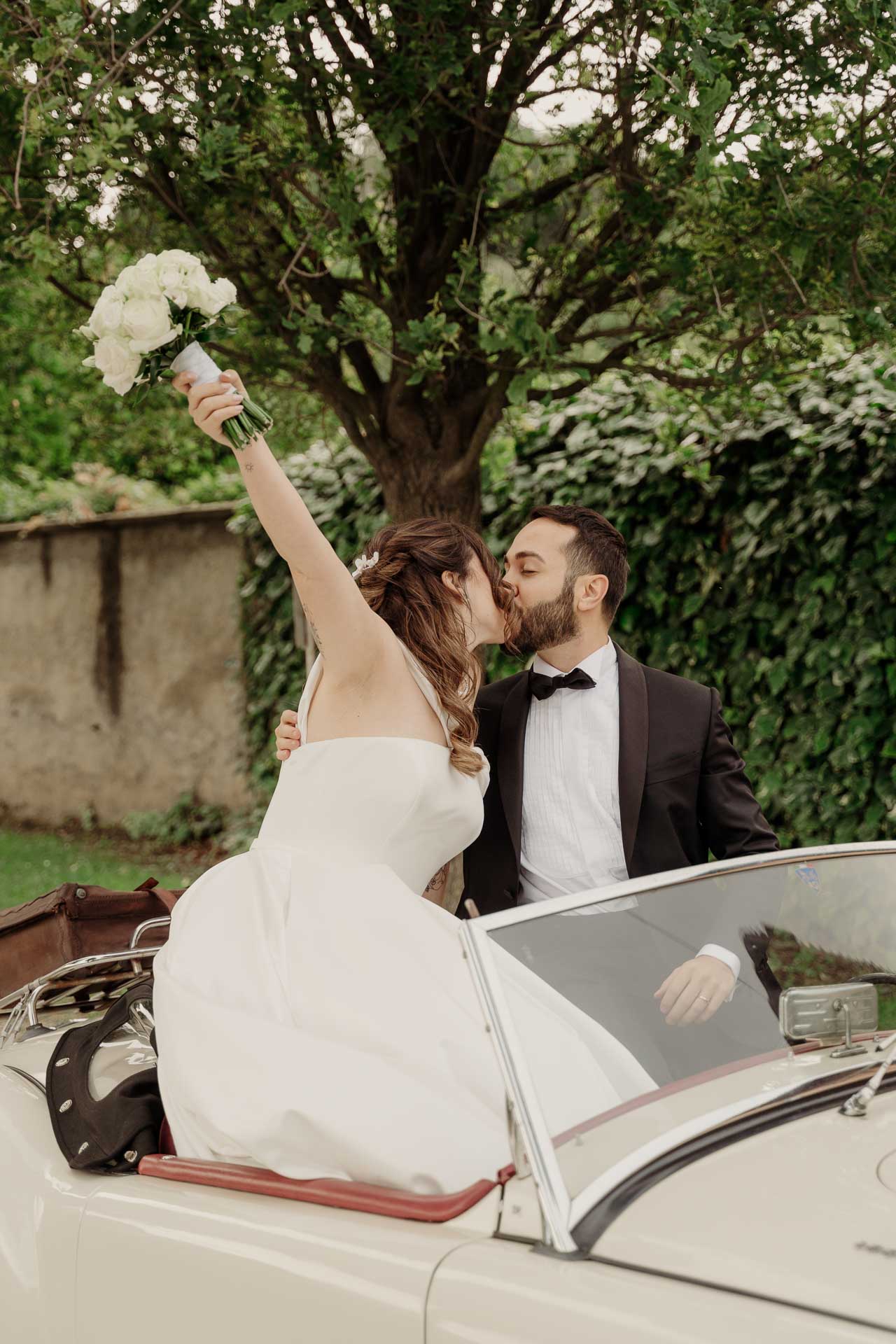 La sposa alza il bouquet e bacia lo sposo sull'auto d'epoca