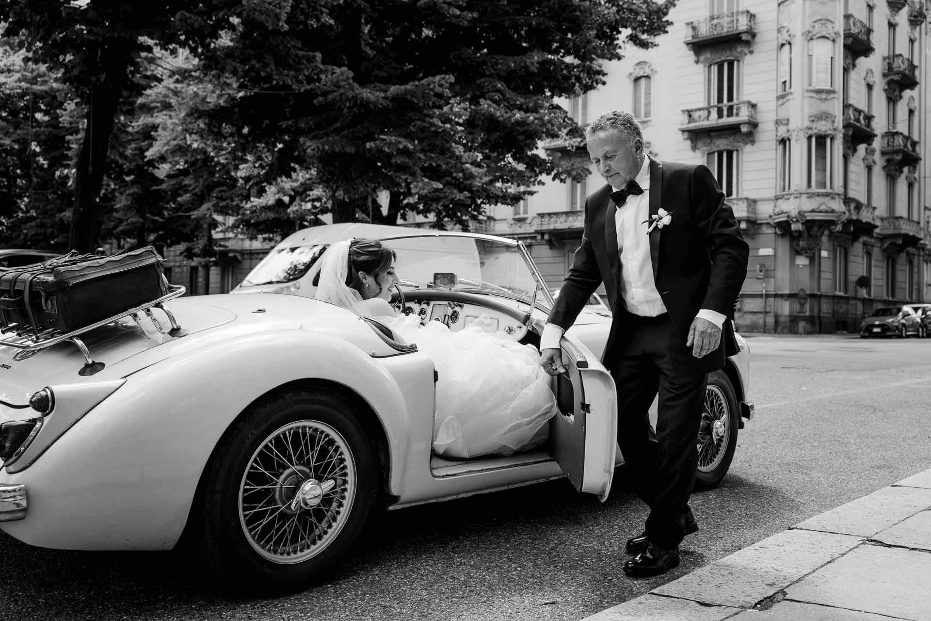 In bianco e nero, un signore aiuta la sposa a scendere dall'auto d'epoca