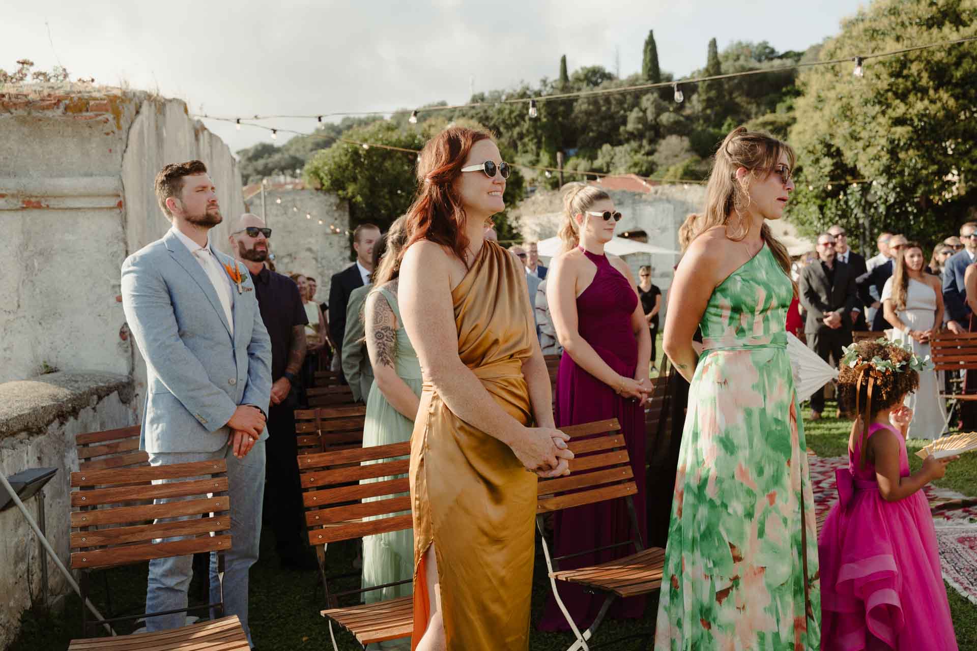 Gli invitati in piedi aspettano la sposa, tra loro una bambina in abito fucsia