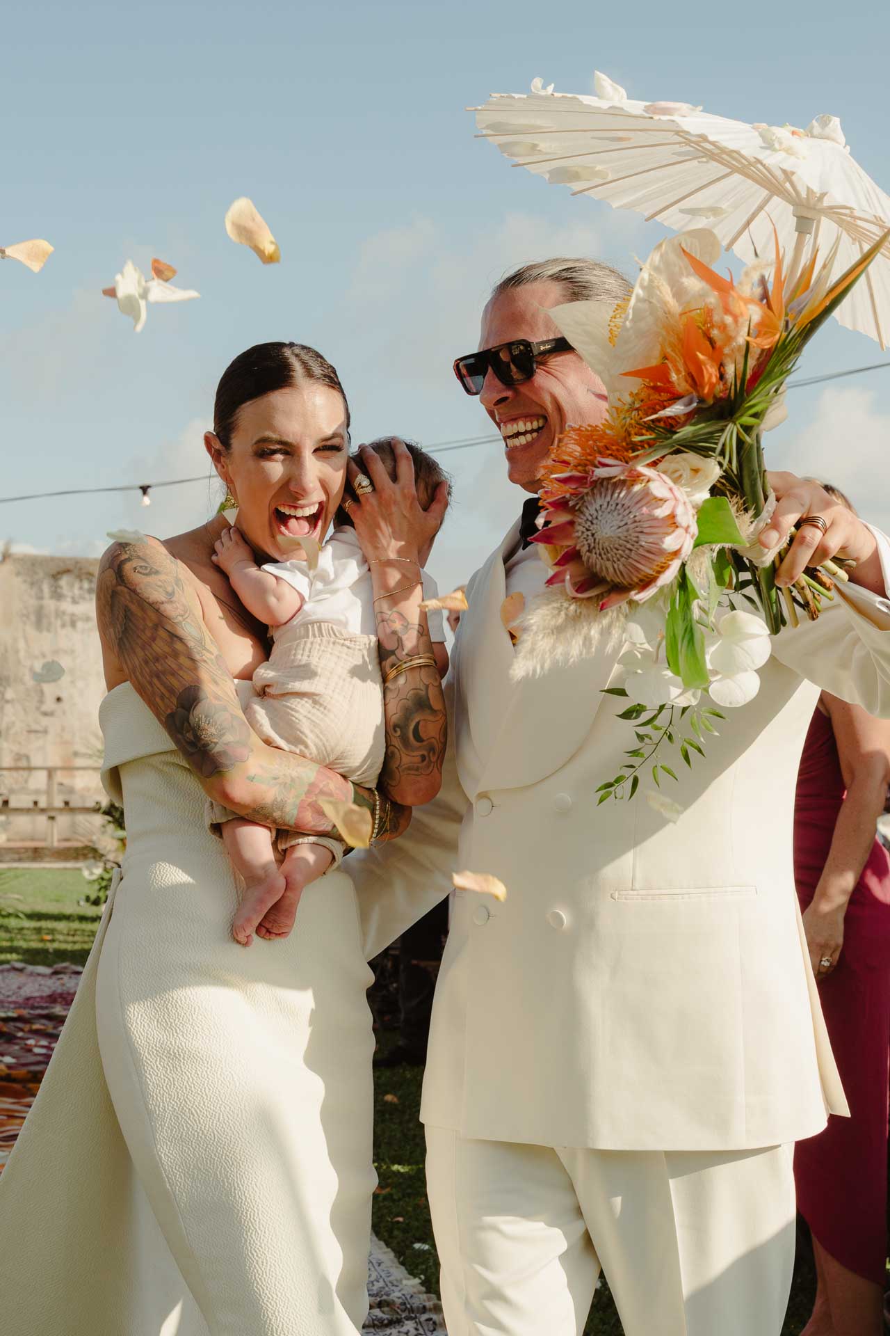 La sposa ride con un bambino in braccio tra i petali, lo sposo con il bouquet e un ombrellino